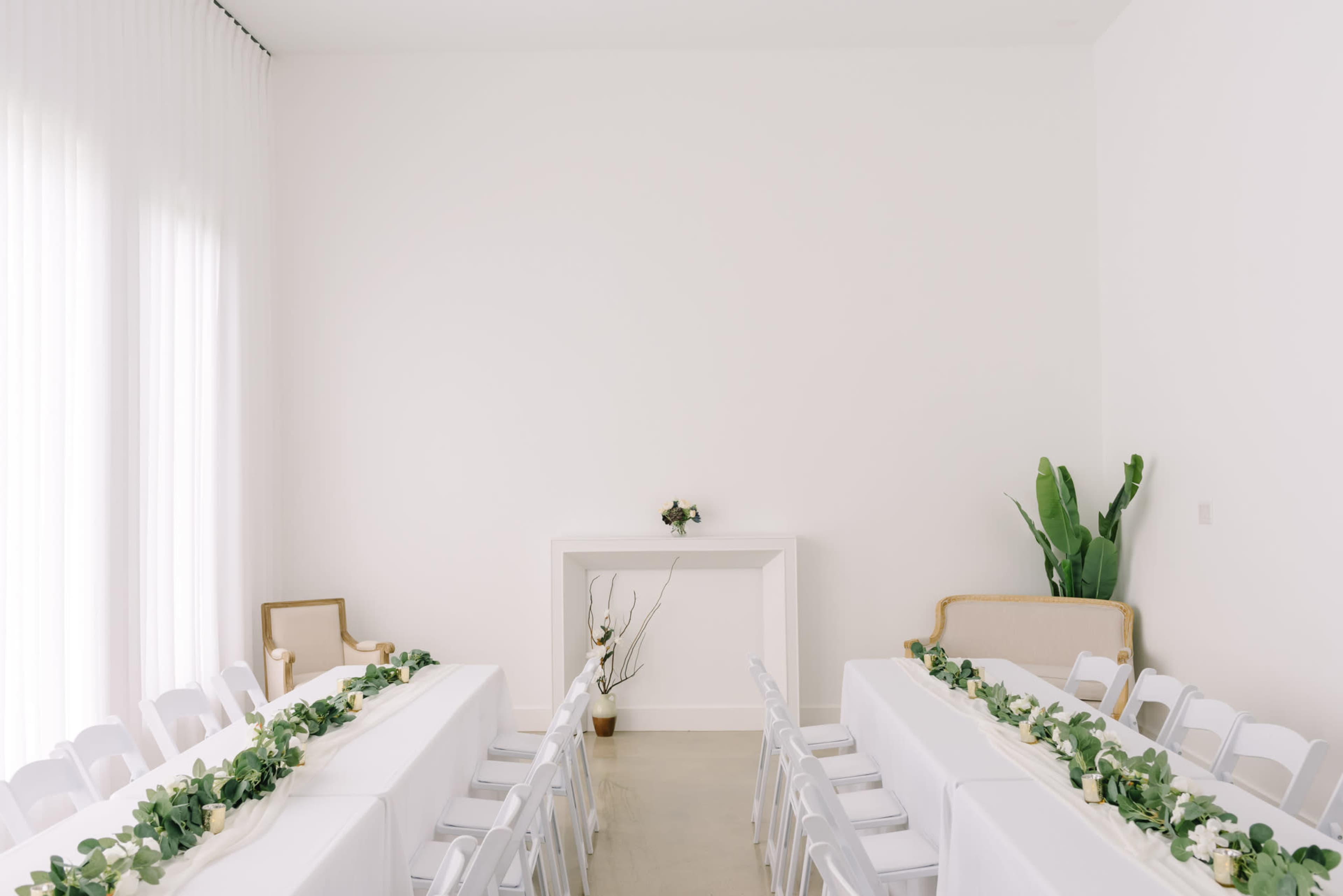 A simple, bright room features two long white tables with chairs arranged on either side, adorned with greenery, and a minimalist white mantel in the background.