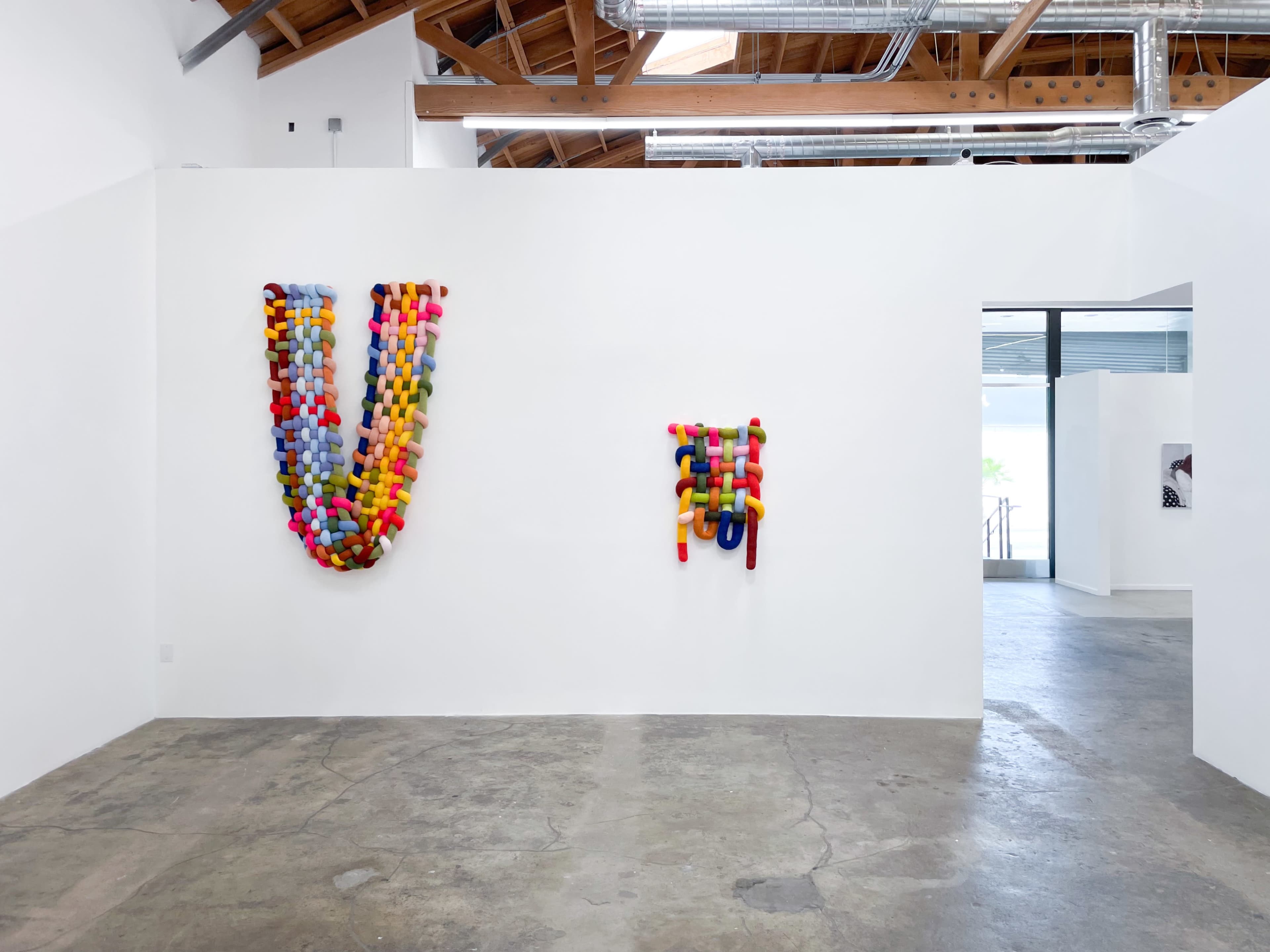 The image shows a modern art gallery featuring colorful textile art pieces on a white wall, with a spacious, minimalistic interior design.