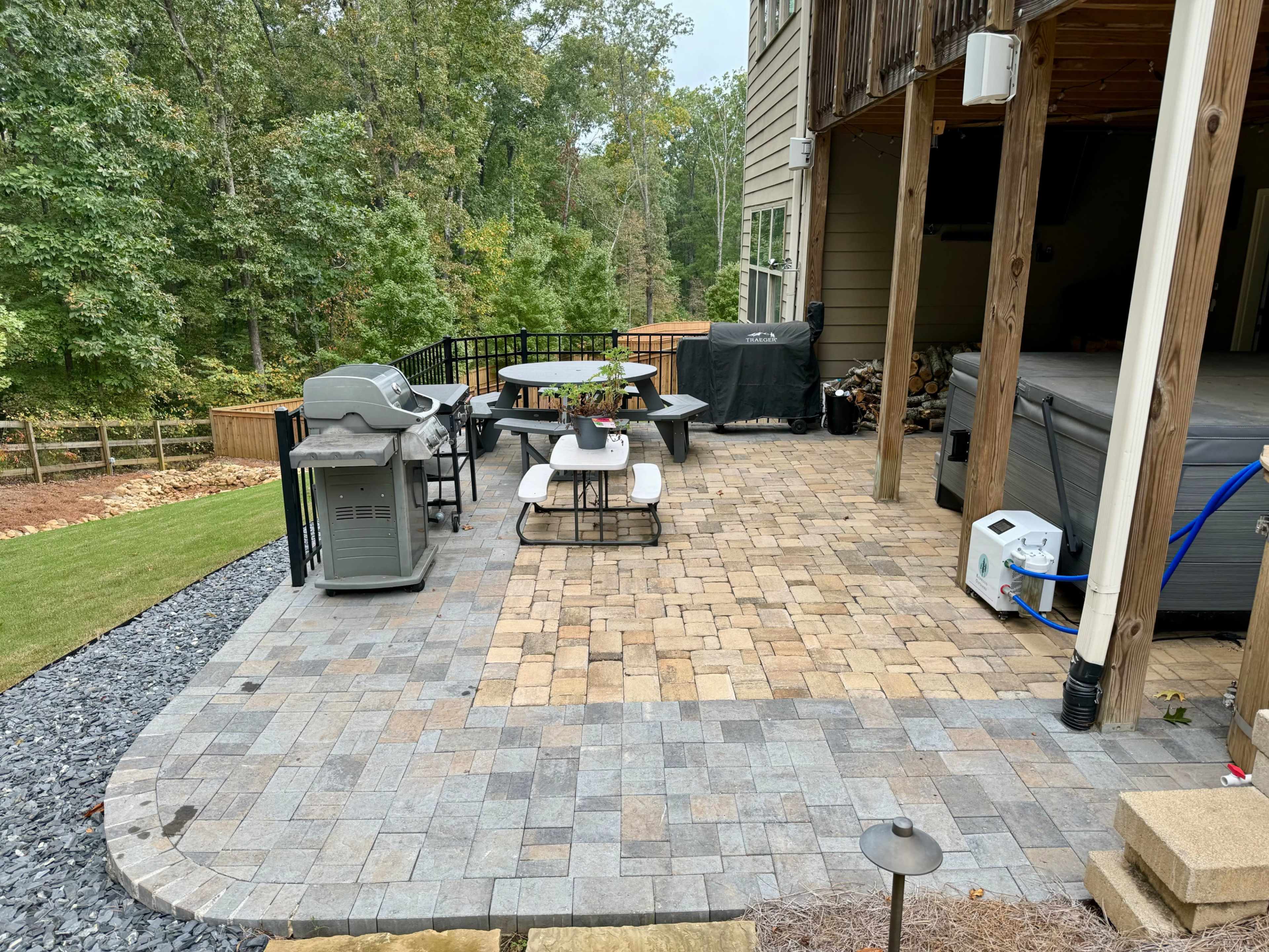 A stone patio area with a table and chairs, a grill, and a hot tub, surrounded by trees and a grassy lawn.