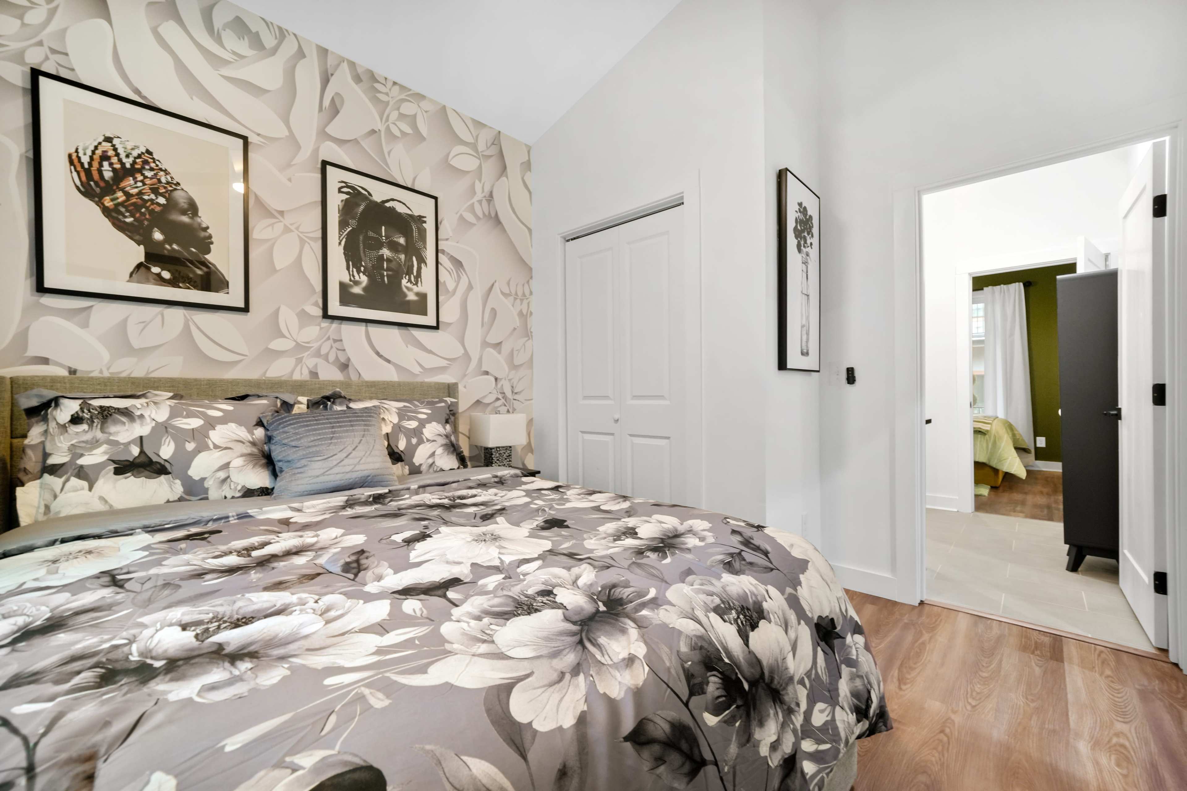 A bedroom features a large floral-patterned bedspread, framed artwork on the wall, and an adjoining door that leads to another room.
