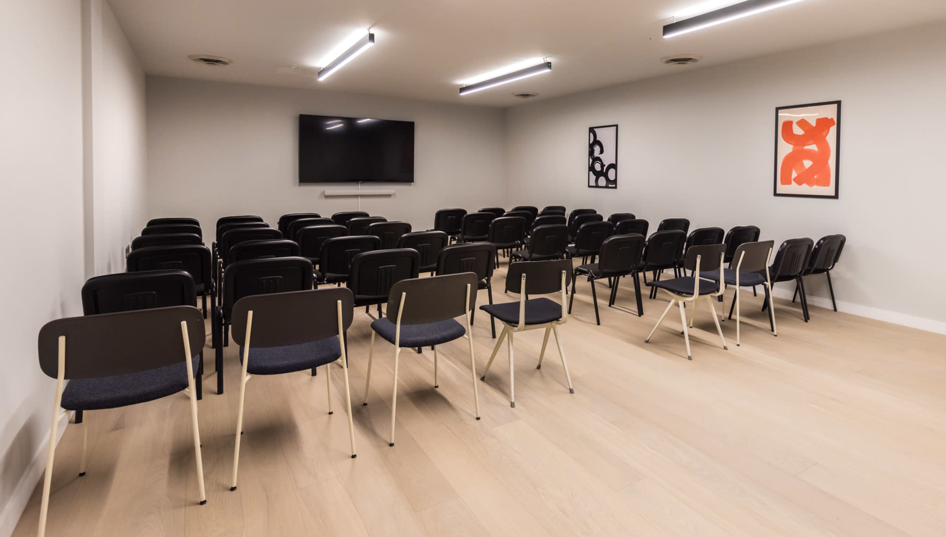 The image shows a conference room with rows of black chairs facing a wall-mounted television, flanked by two abstract art pieces.