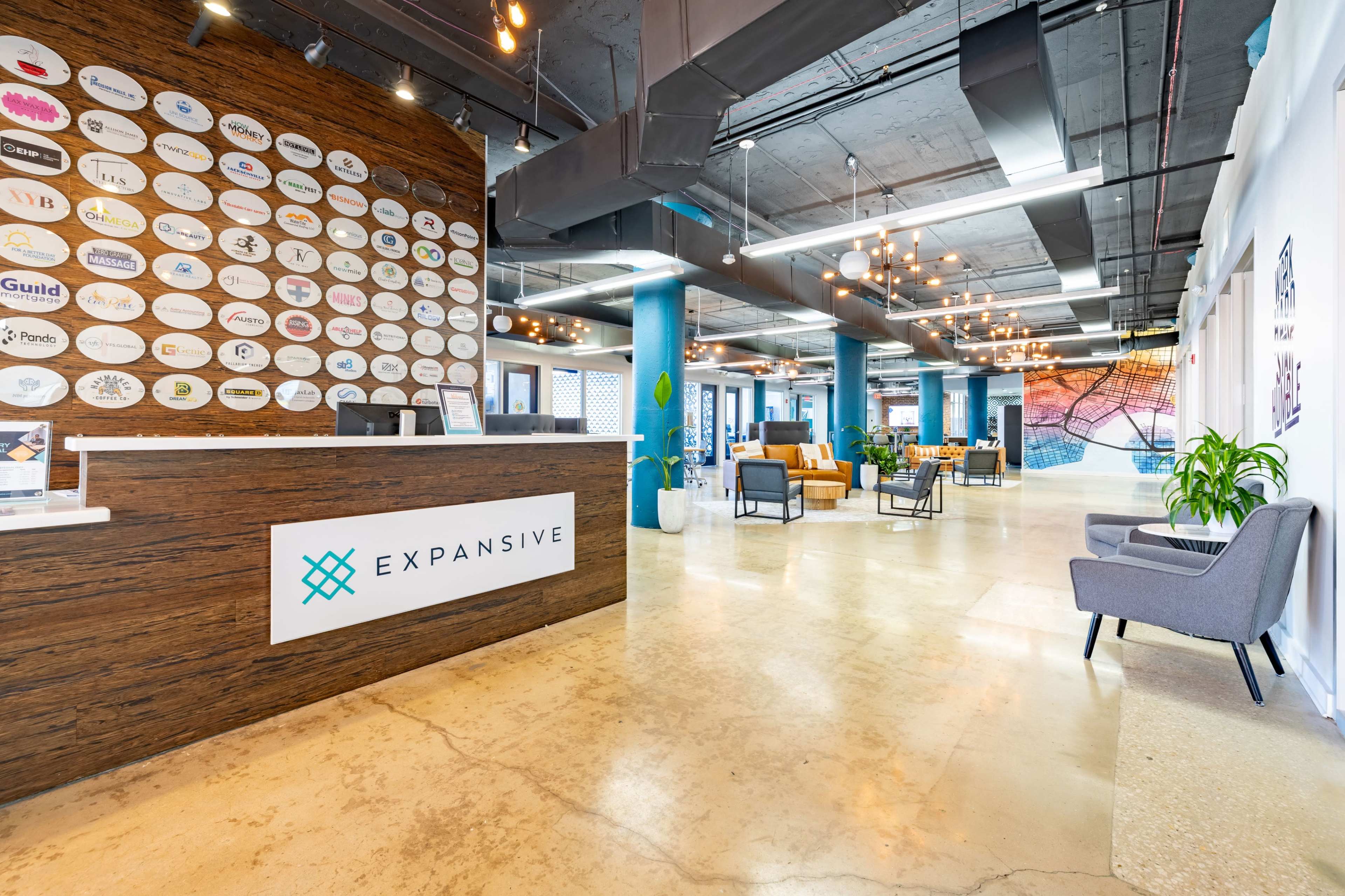 The image shows a modern office reception area with a wooden front desk displaying the name "EXPANSIVE," surrounded by colorful seating and decorative wall art.