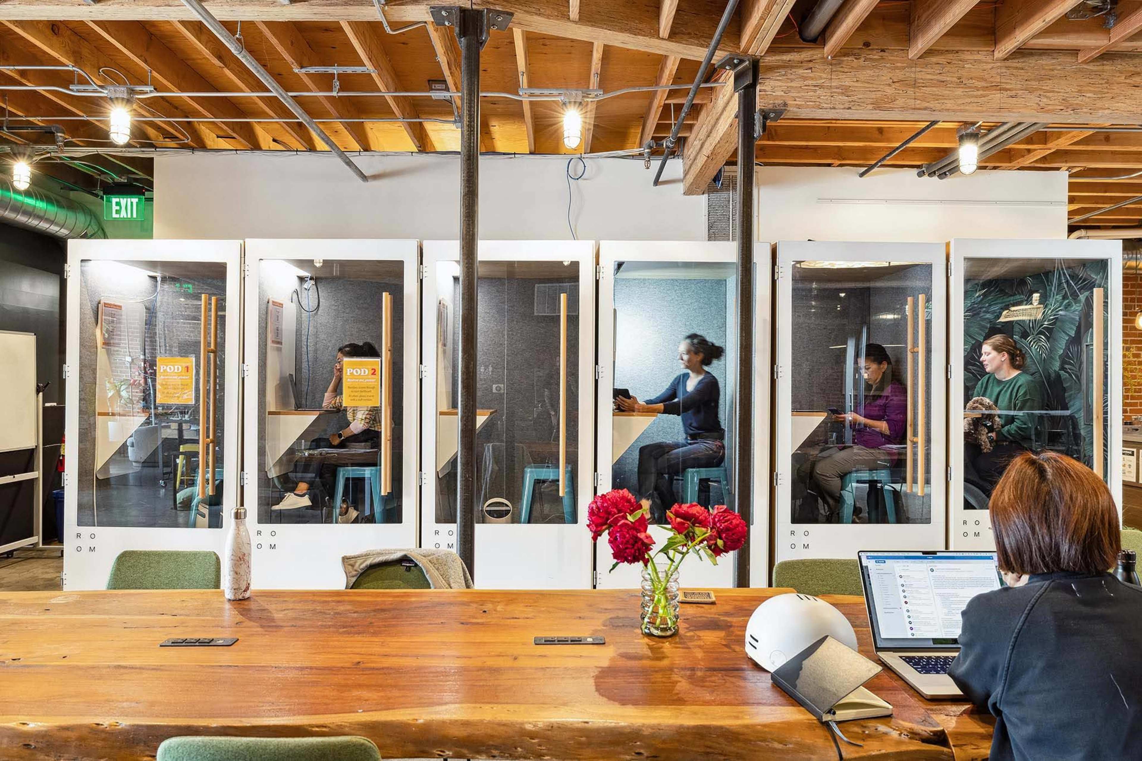 A modern workspace featuring soundproof booths for individual use, with three people inside and a communal table in the foreground.