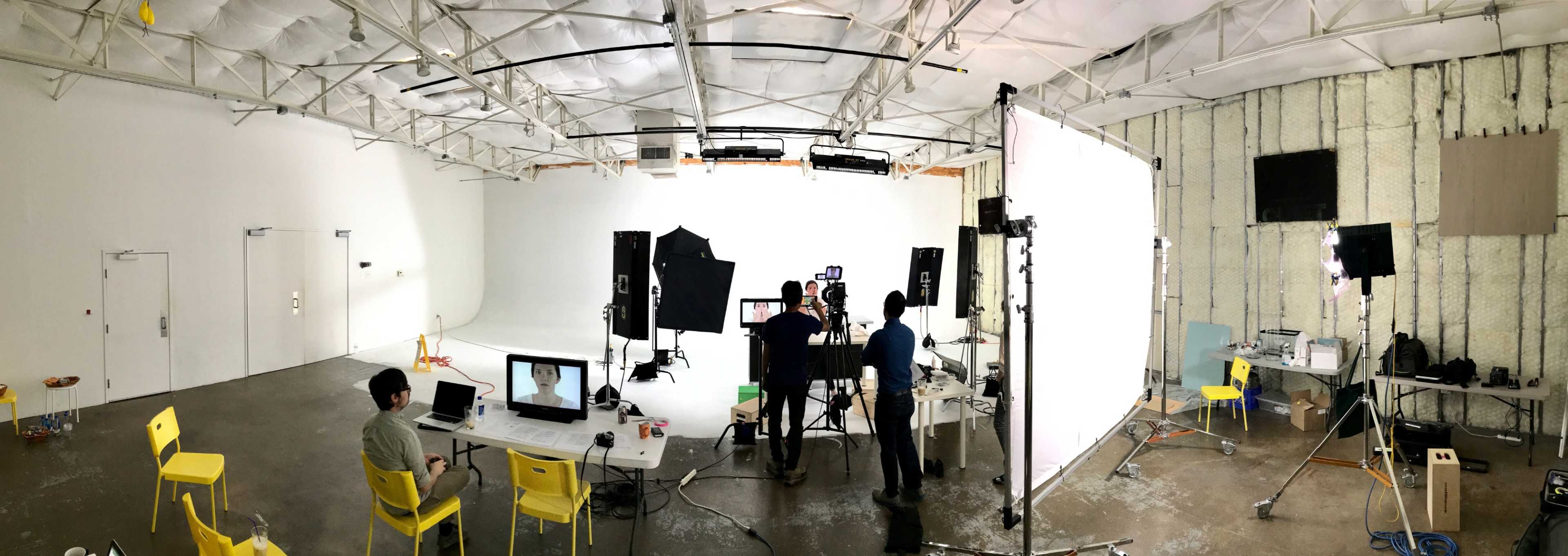 A photo shows a spacious filming studio with lighting equipment, cameras, and a production team working on a set surrounded by various chairs and tables.