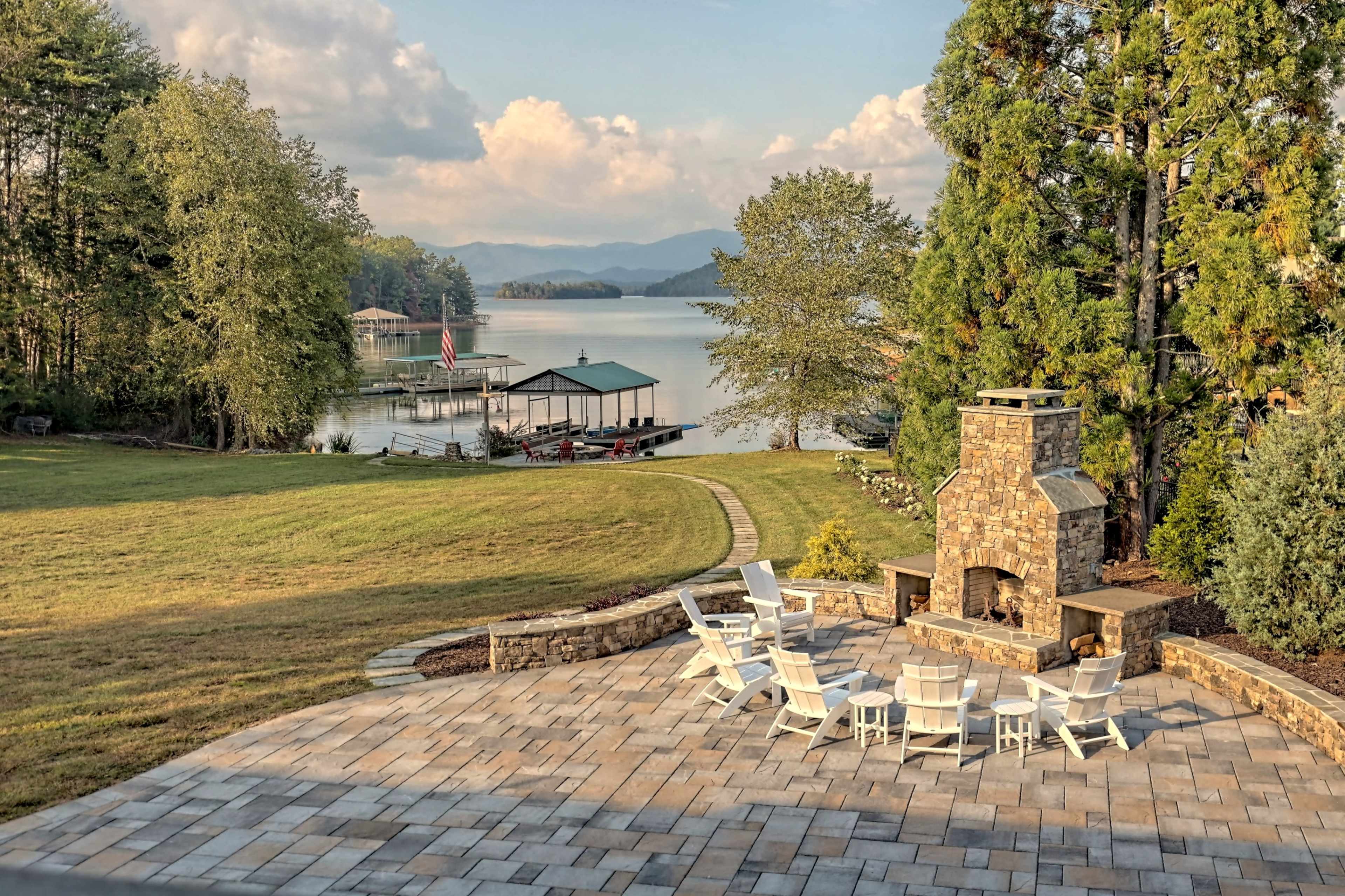 A lakeside patio with white chairs arranged around a stone fireplace, overlooking a calm lake and distant mountains.