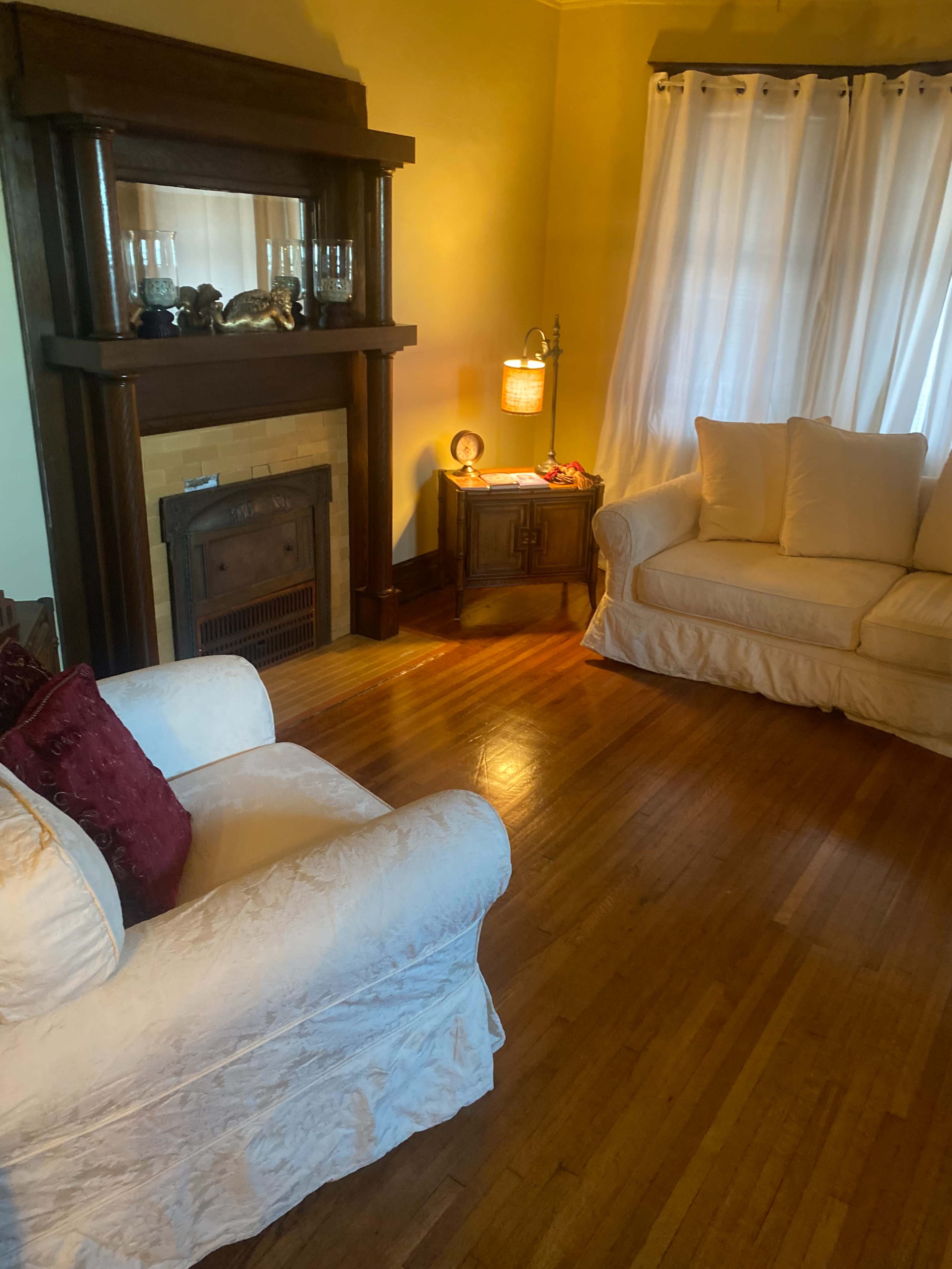 The image shows a cozy living room featuring a light-colored couch, a fireplace with decorative items, and a lamp beside a small table, illuminated by natural light from a window with white curtains.