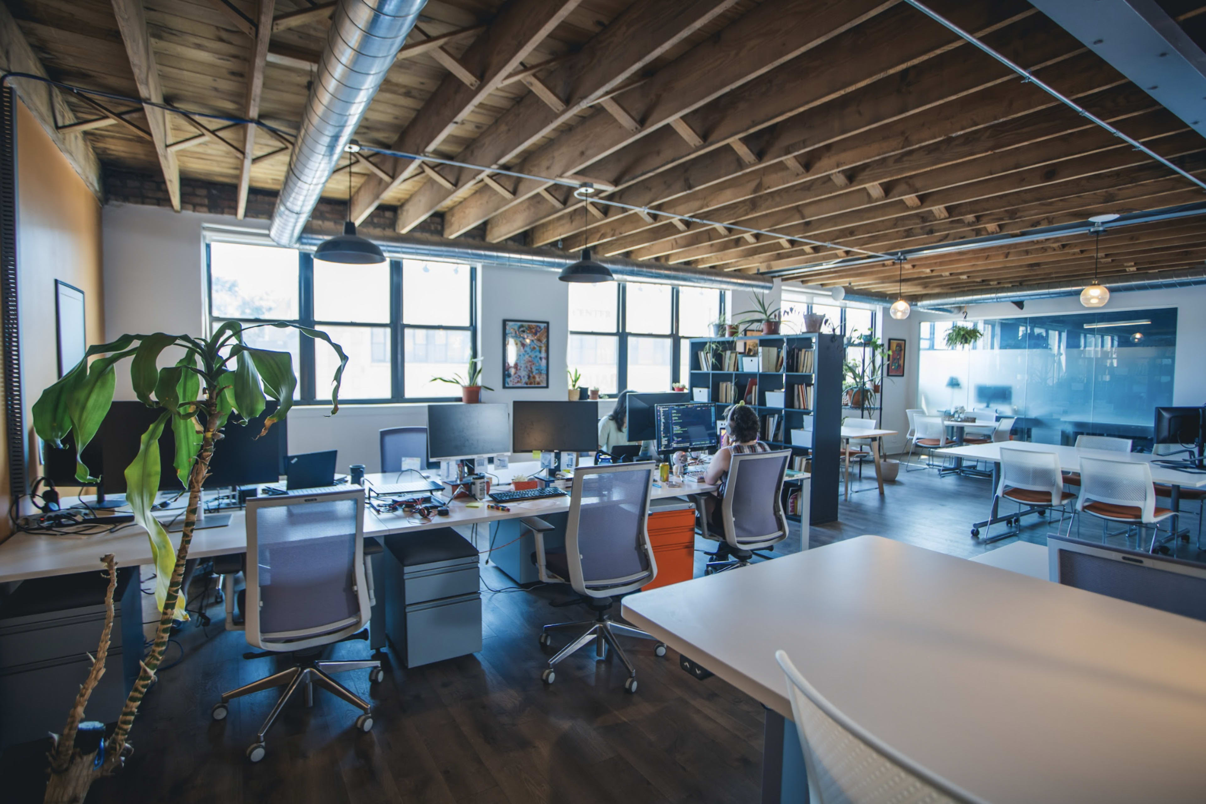 A modern open-plan office space with multiple desks, computer monitors, and large windows allowing natural light.