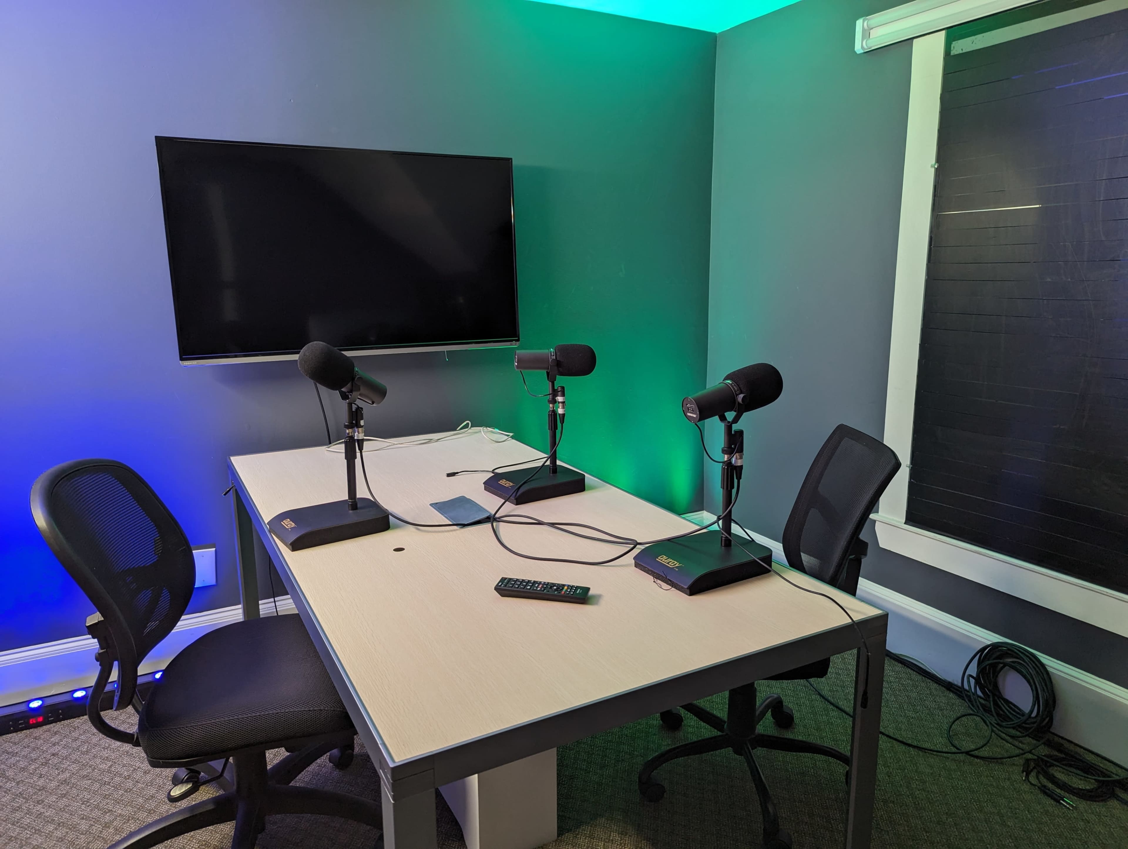 A recording studio with a table set for four microphones and two chairs, featuring a television screen on the wall and green ambient lighting.