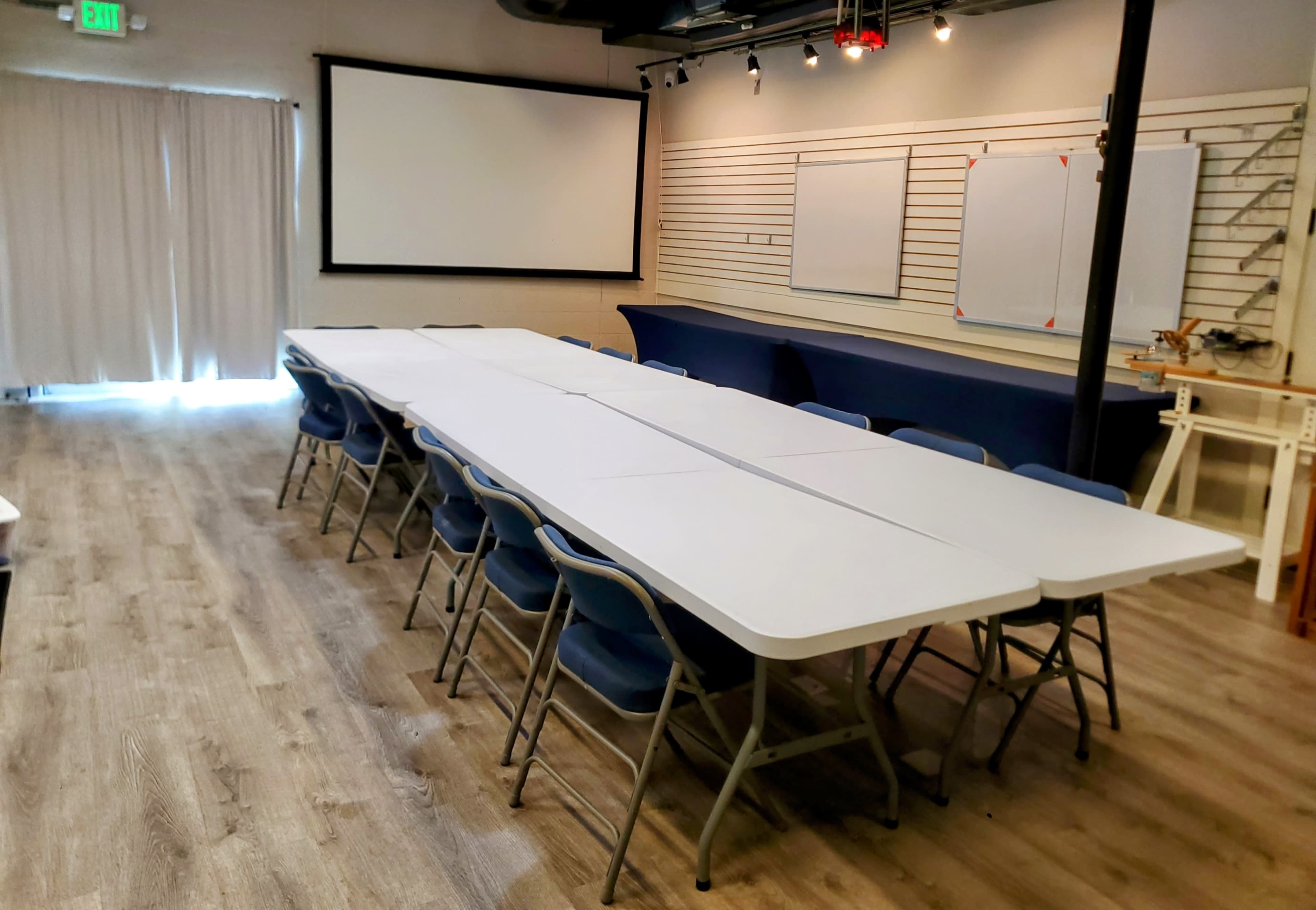 The image shows a conference room featuring a long white table surrounded by folding chairs, with a projector screen and blank walls suitable for presentations.
