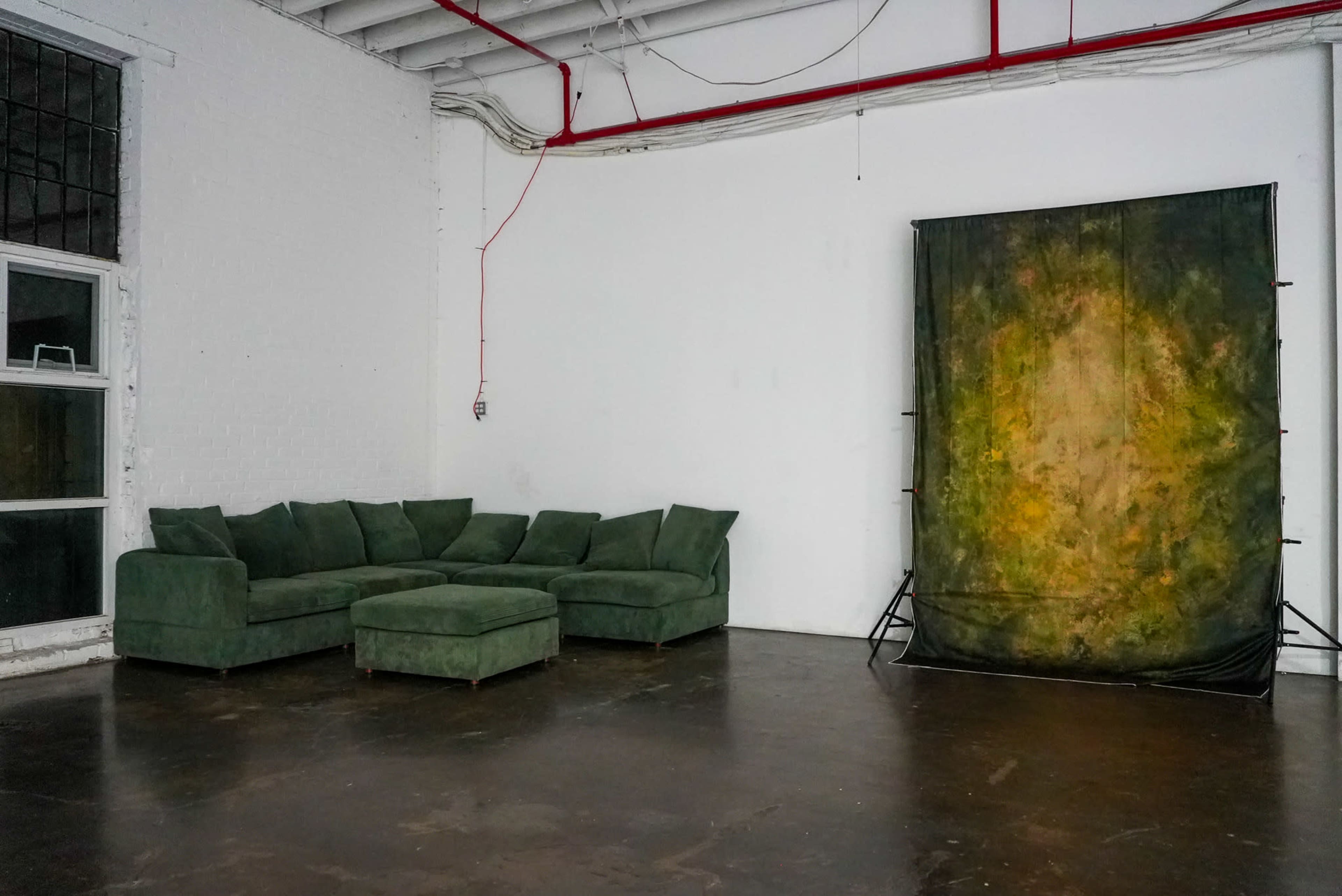 The image shows a spacious, empty room with a green sectional sofa on the left and a backdrop stand with a textured, multicolored fabric on the right.