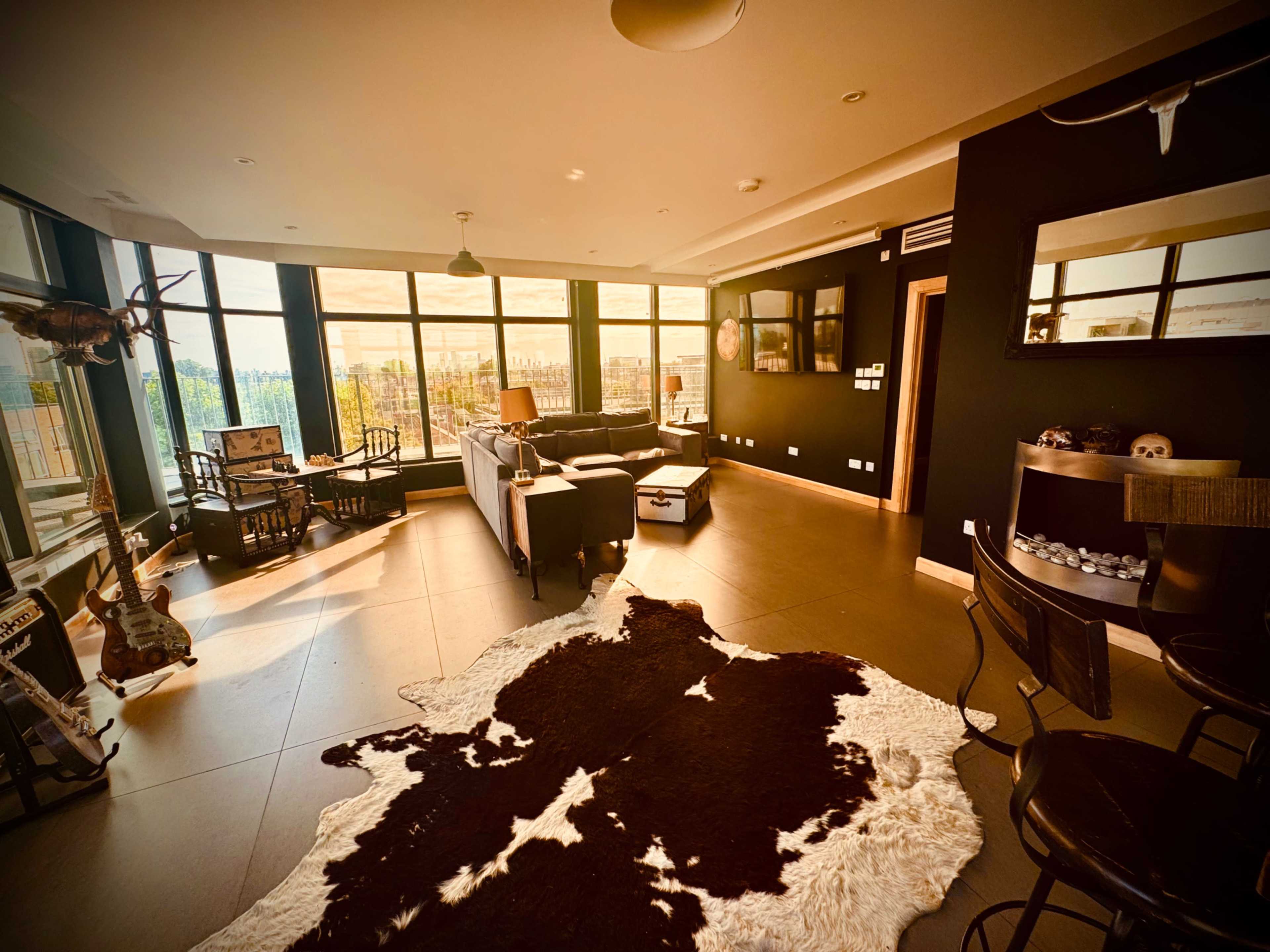 The image shows a spacious living room with large windows, modern furniture, and a patterned rug on the floor.