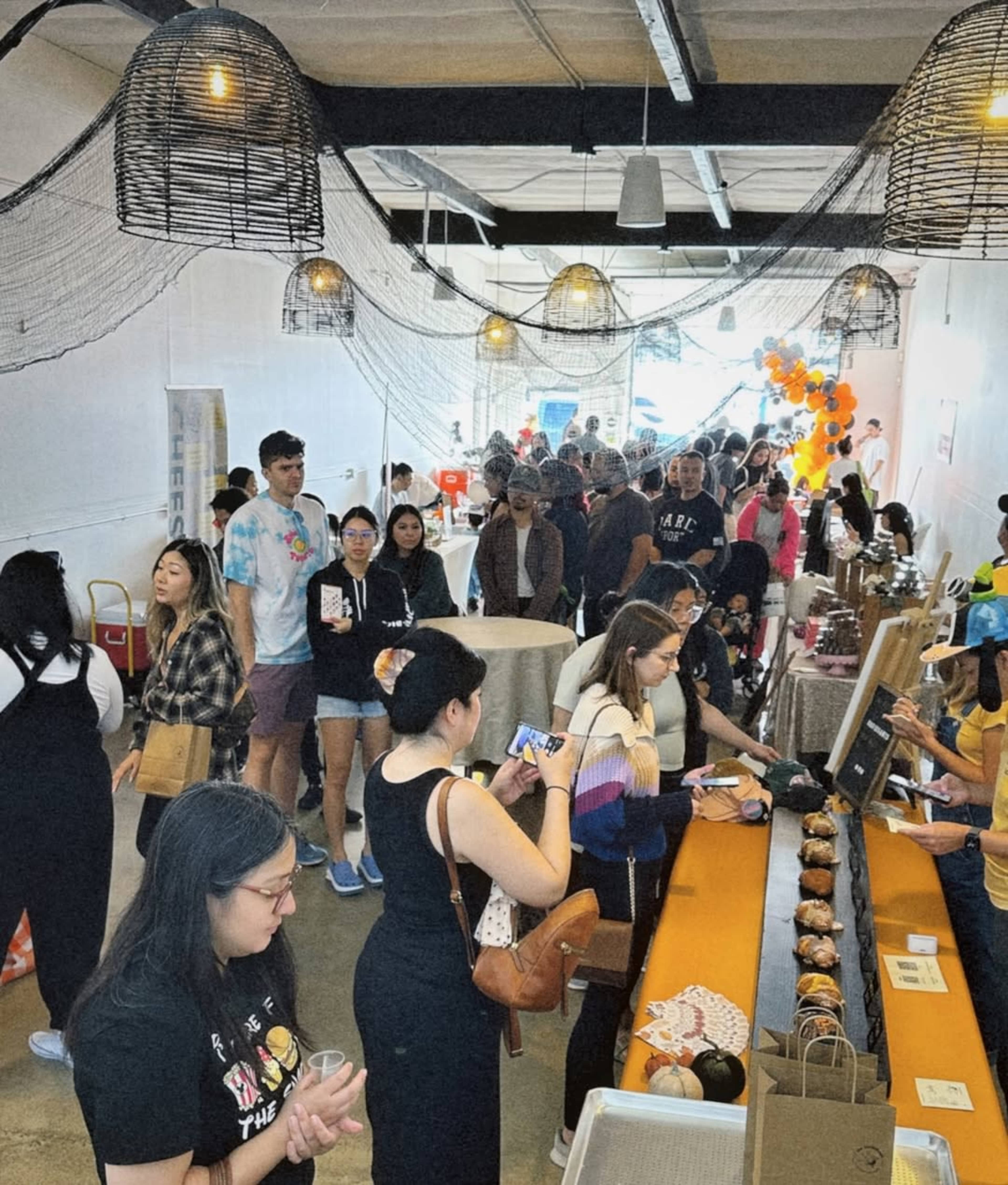 A large crowd of people gathers inside a spacious venue filled with various booths and tables, while decorations hang from the ceiling.