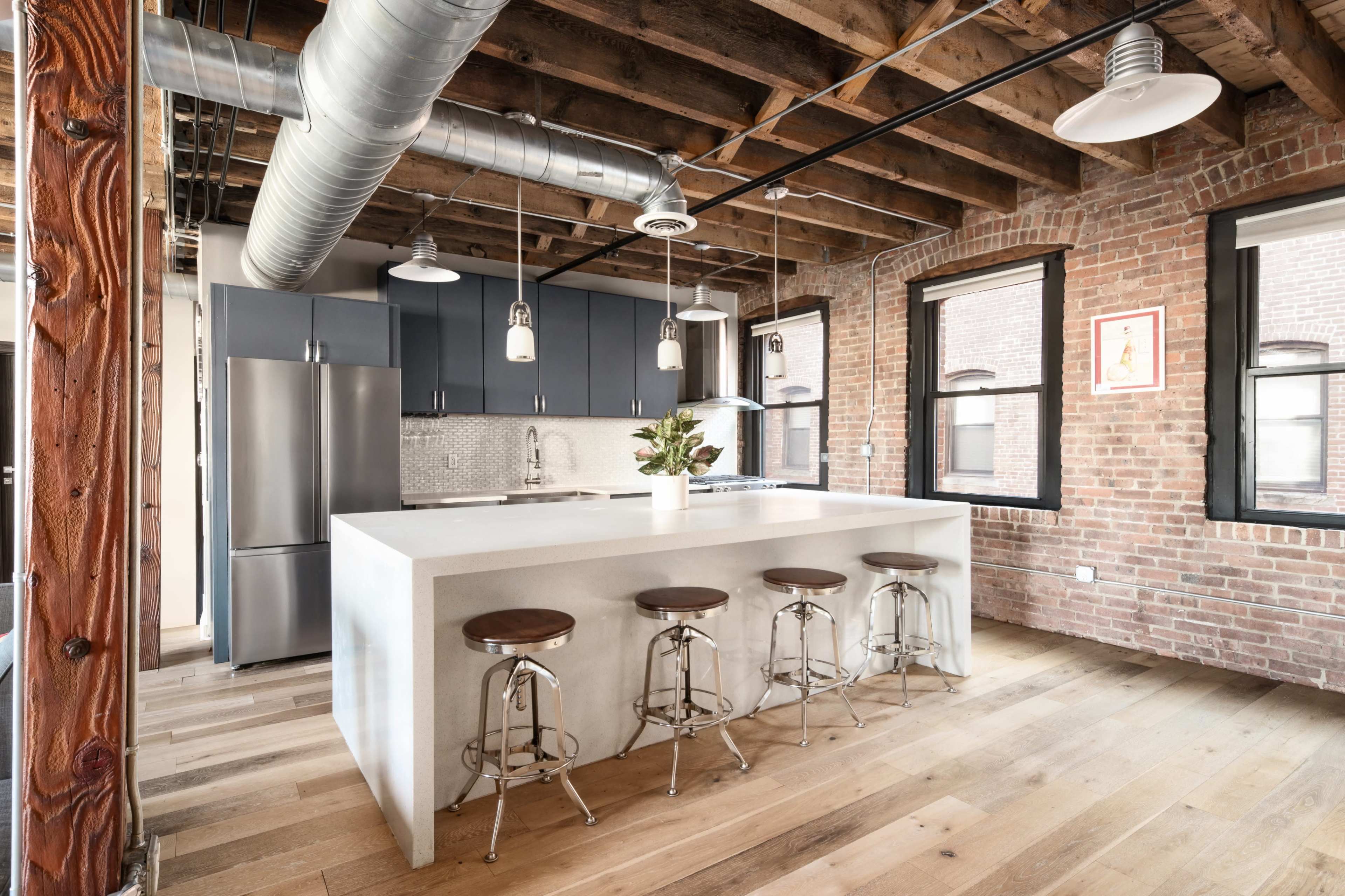 The image shows a modern kitchen with exposed brick walls, wooden flooring, and a large island with four metal stools.
