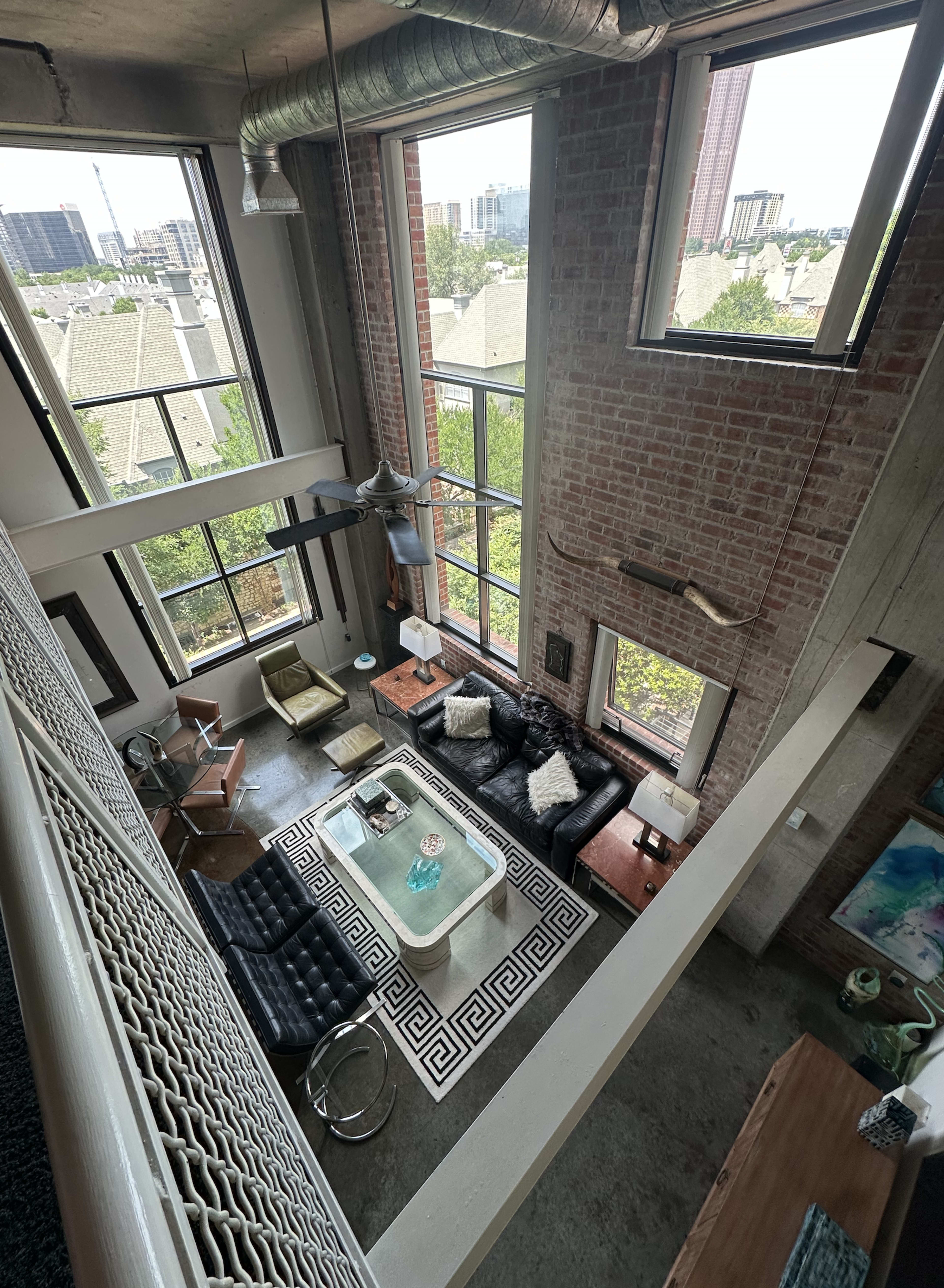 The image shows a modern loft-style living room with large windows, exposed brick walls, and a high ceiling.