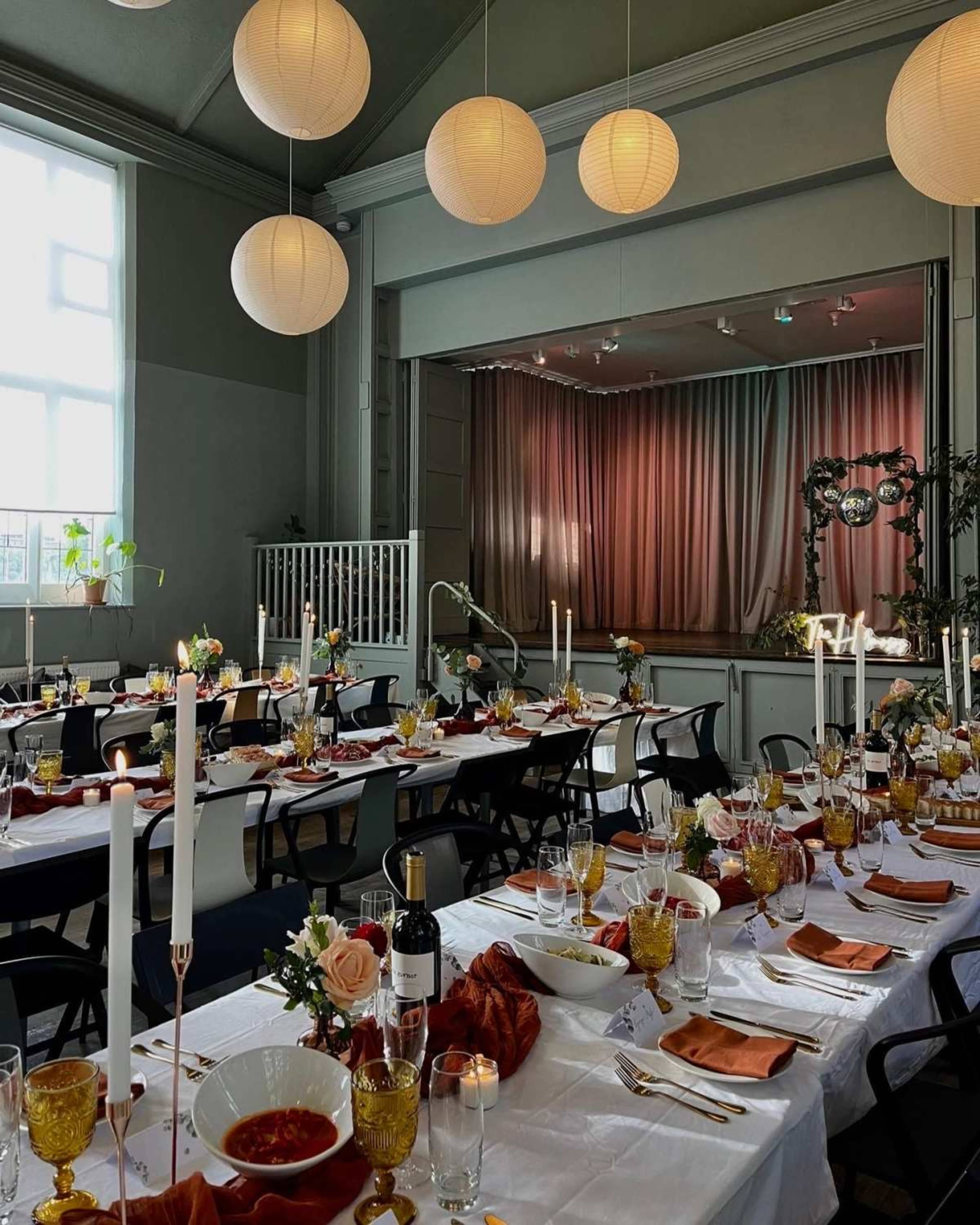 A beautifully arranged dining hall features tables set with elegant tableware, flickering candles, and floral centerpieces, with soft lantern lights overhead and a stage visible in the background.