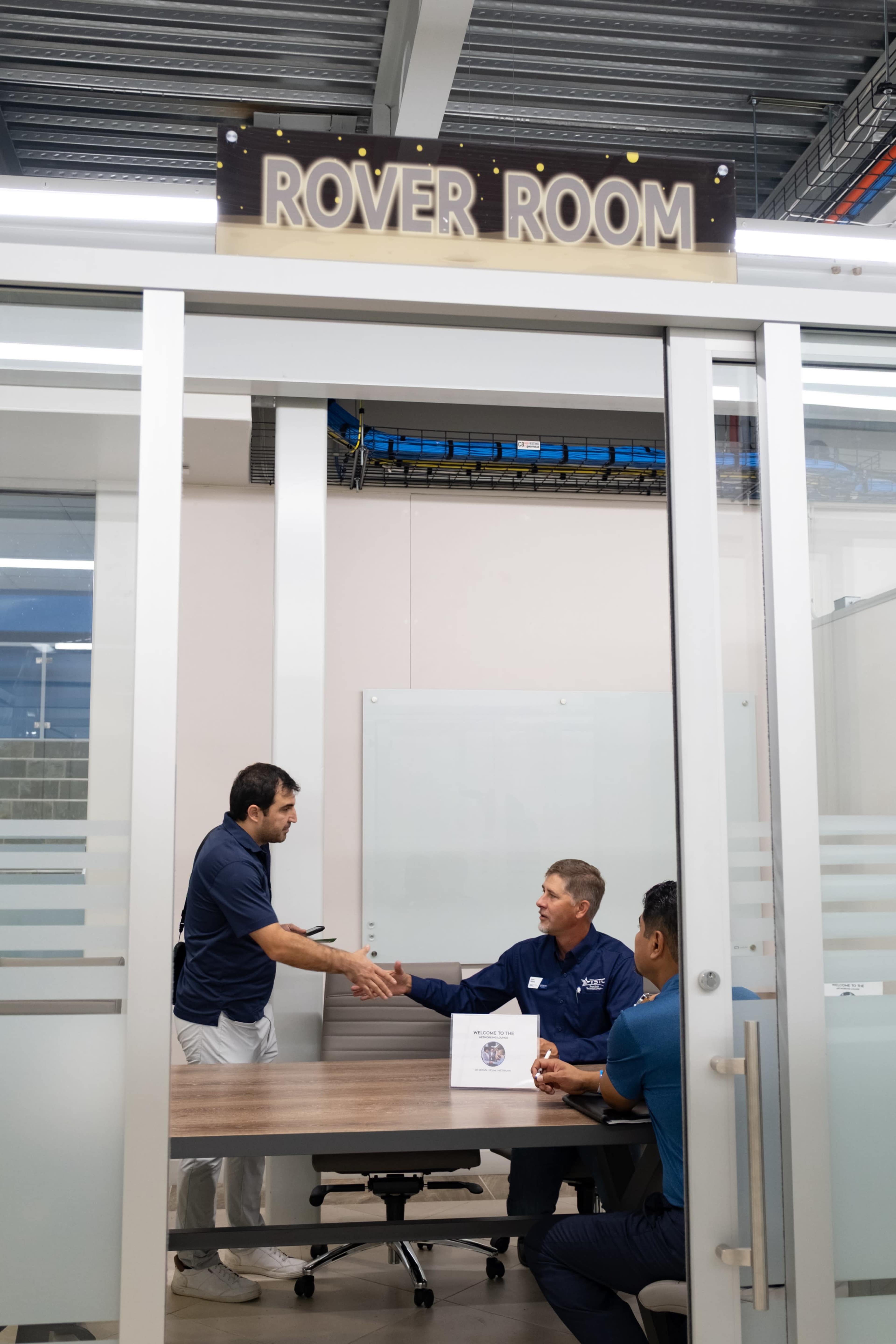Two men are shaking hands in a glass-walled conference room labeled "ROVER ROOM," while a third man sits at the table with a box.