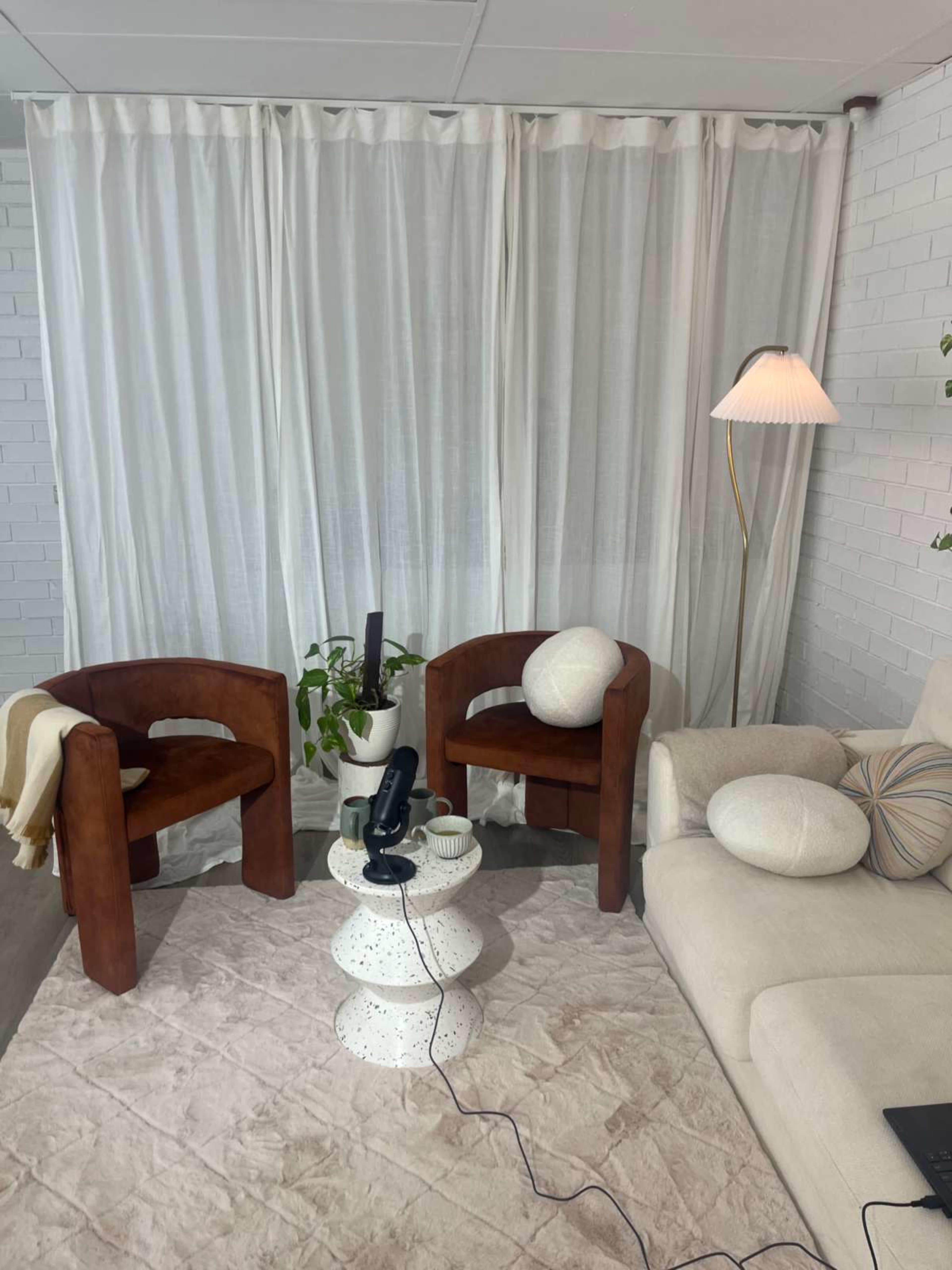The image shows a cozy living room setting with two wooden chairs, a small round table, and a light-colored sofa, all arranged near a curtain and potted plant.