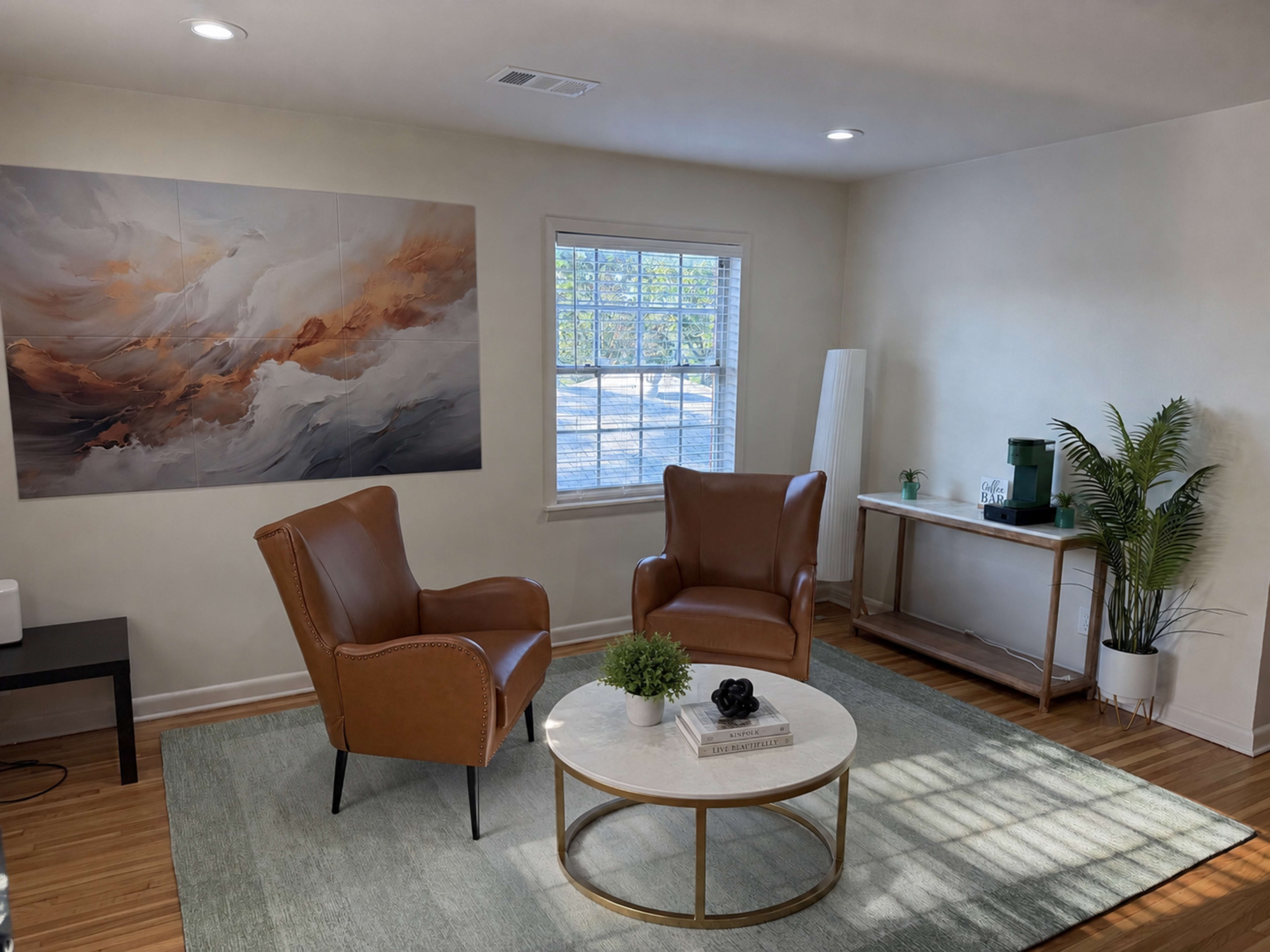 The image shows a bright, modern living room featuring two brown leather chairs, a round coffee table, and a large abstract painting on the wall.