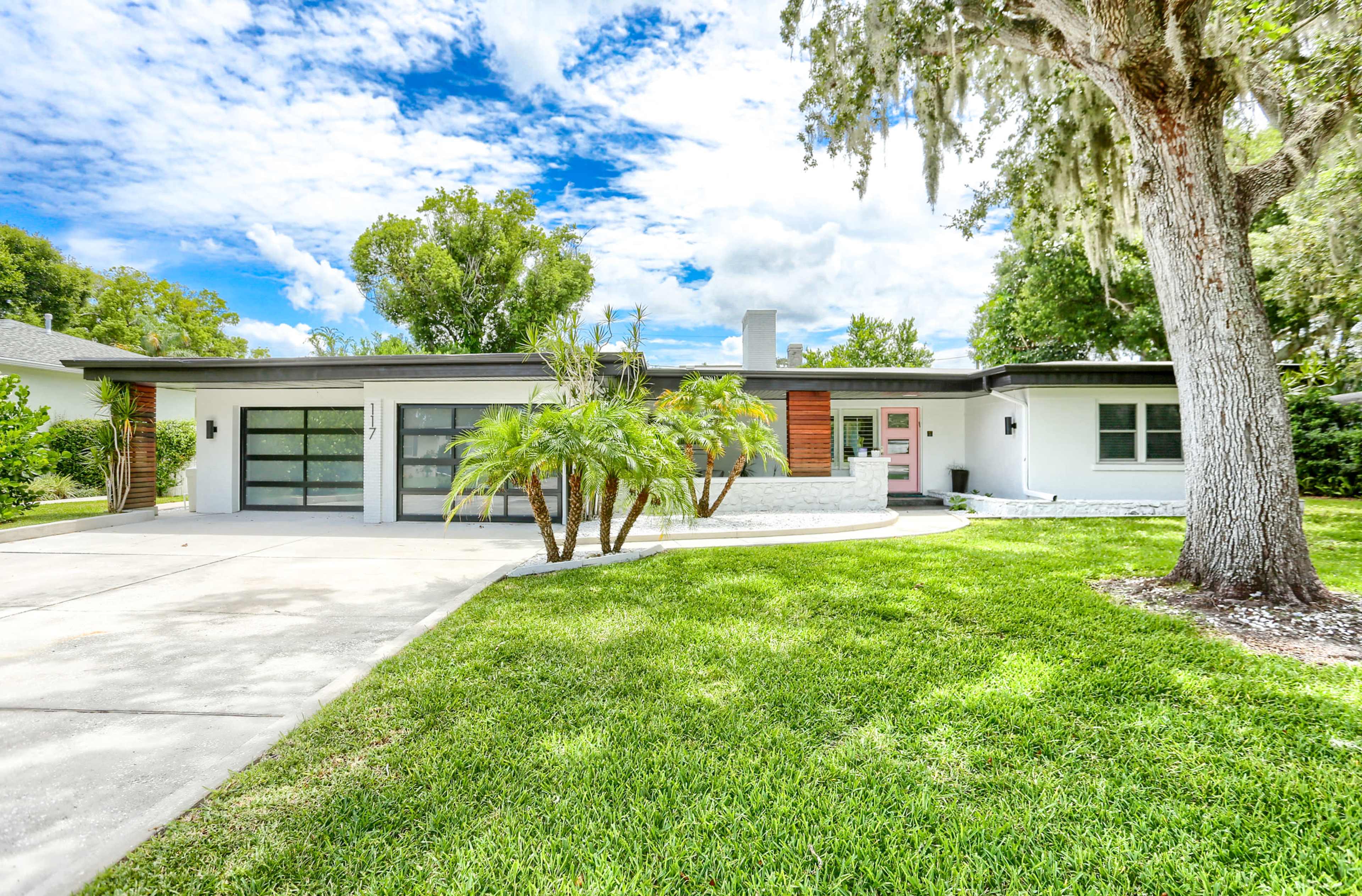 A modern single-story house features a landscaped front yard with palm trees and a driveway leading to a glass garage door.