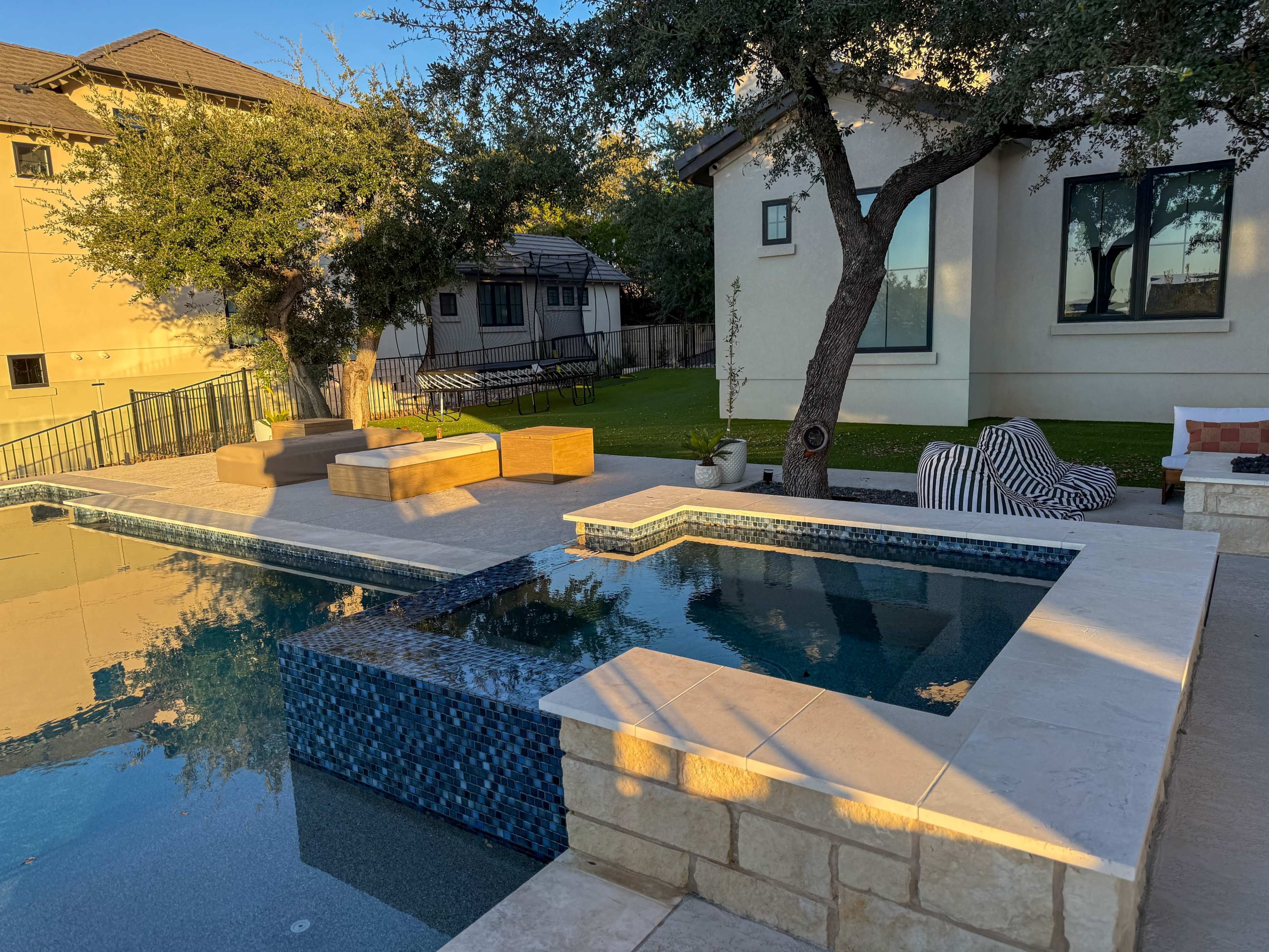 A modern pool area features a stone wall, wooden benches, and an adjacent grassy yard, framed by residential buildings and trees.