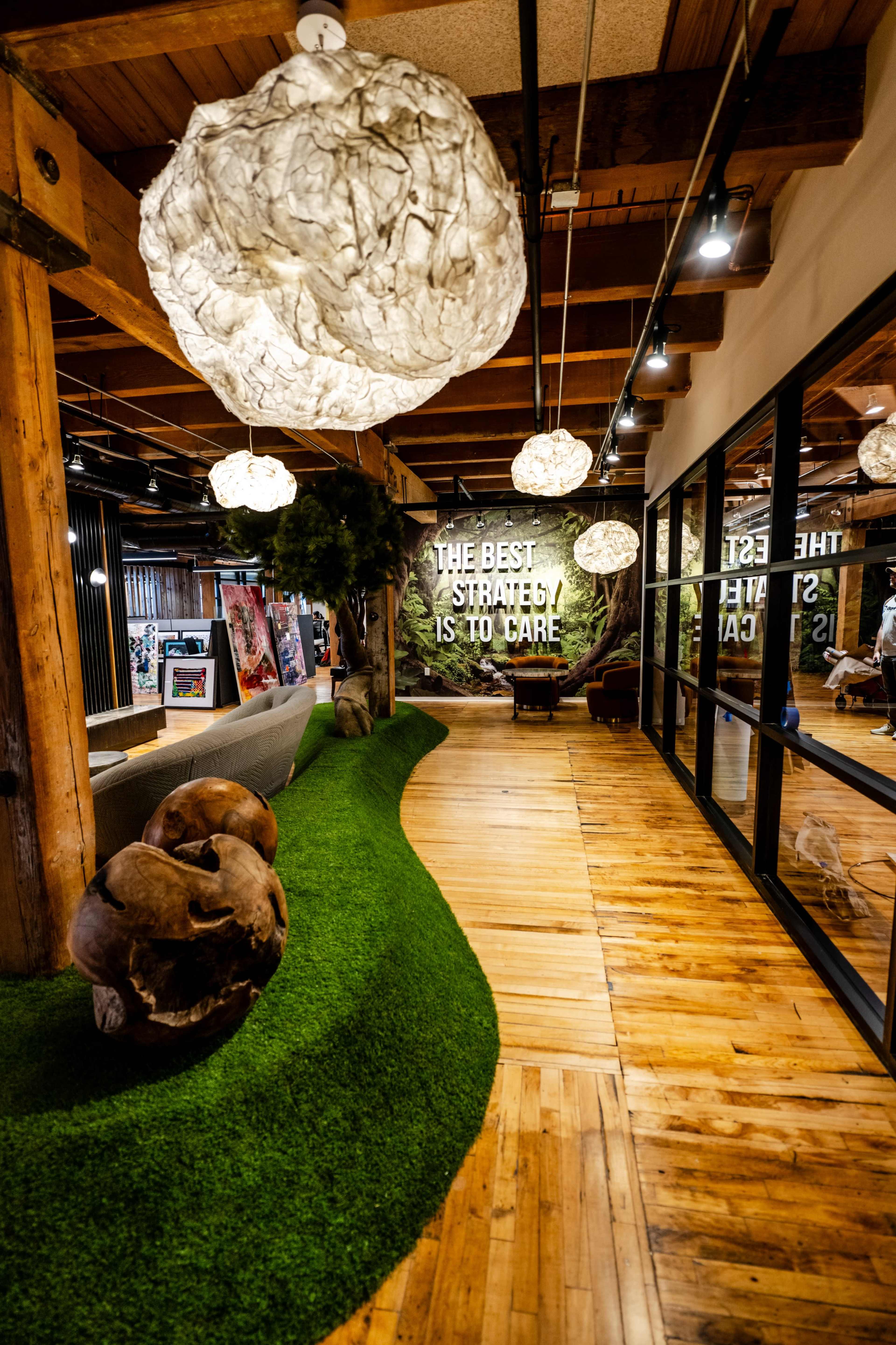 The image shows an interior space featuring a path of artificial grass, wooden flooring, and decorative lighting, with a wall displaying the phrase "THE BEST STRATEGY IS TO CARE."