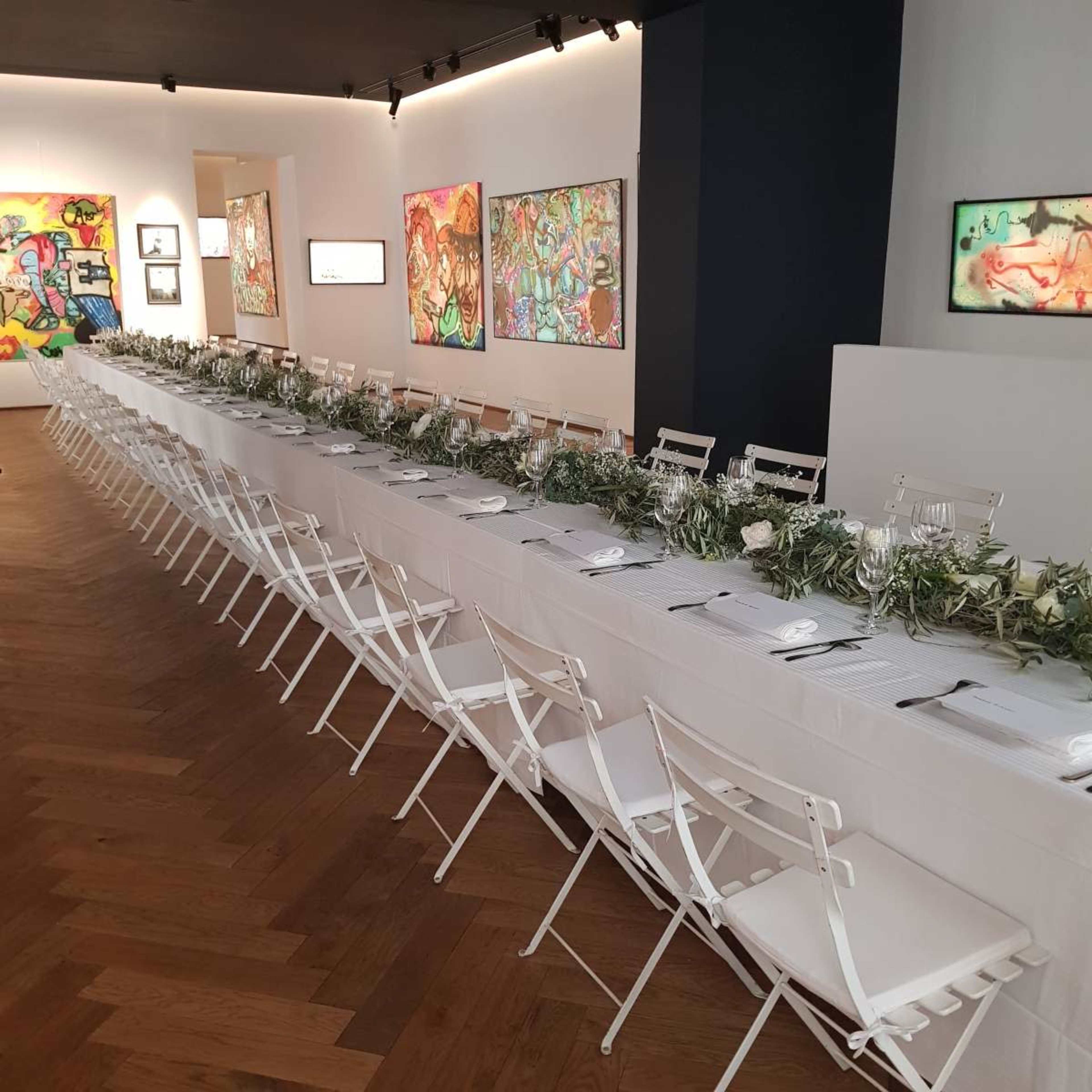 A long dining table is set with white tablecloths and surrounded by white chairs, decorated with greenery, in a gallery featuring colorful artwork on the walls.
