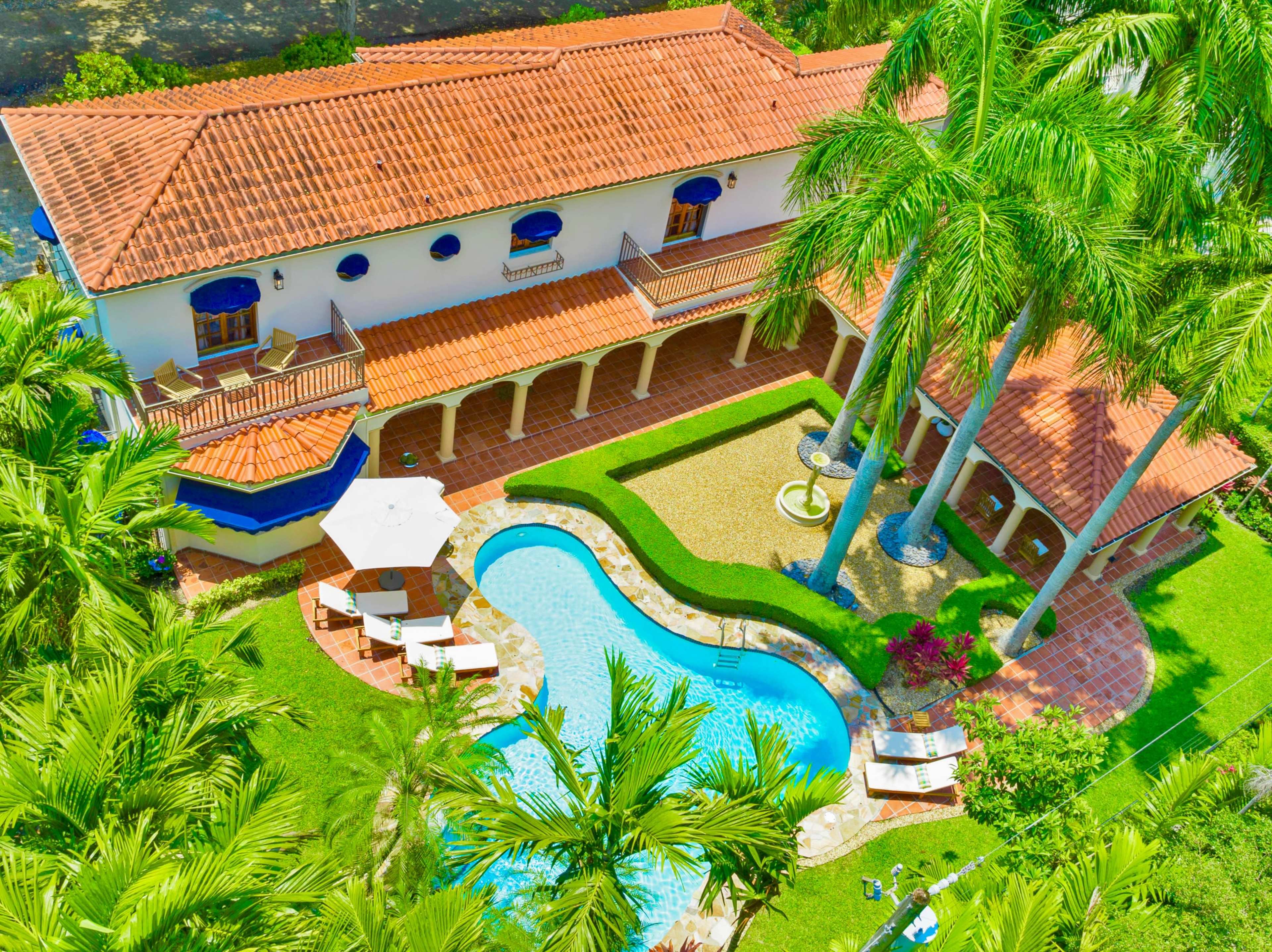 An aerial view of a large house with a red-tiled roof, surrounded by palm trees and featuring a swimming pool and a fountain in the backyard.