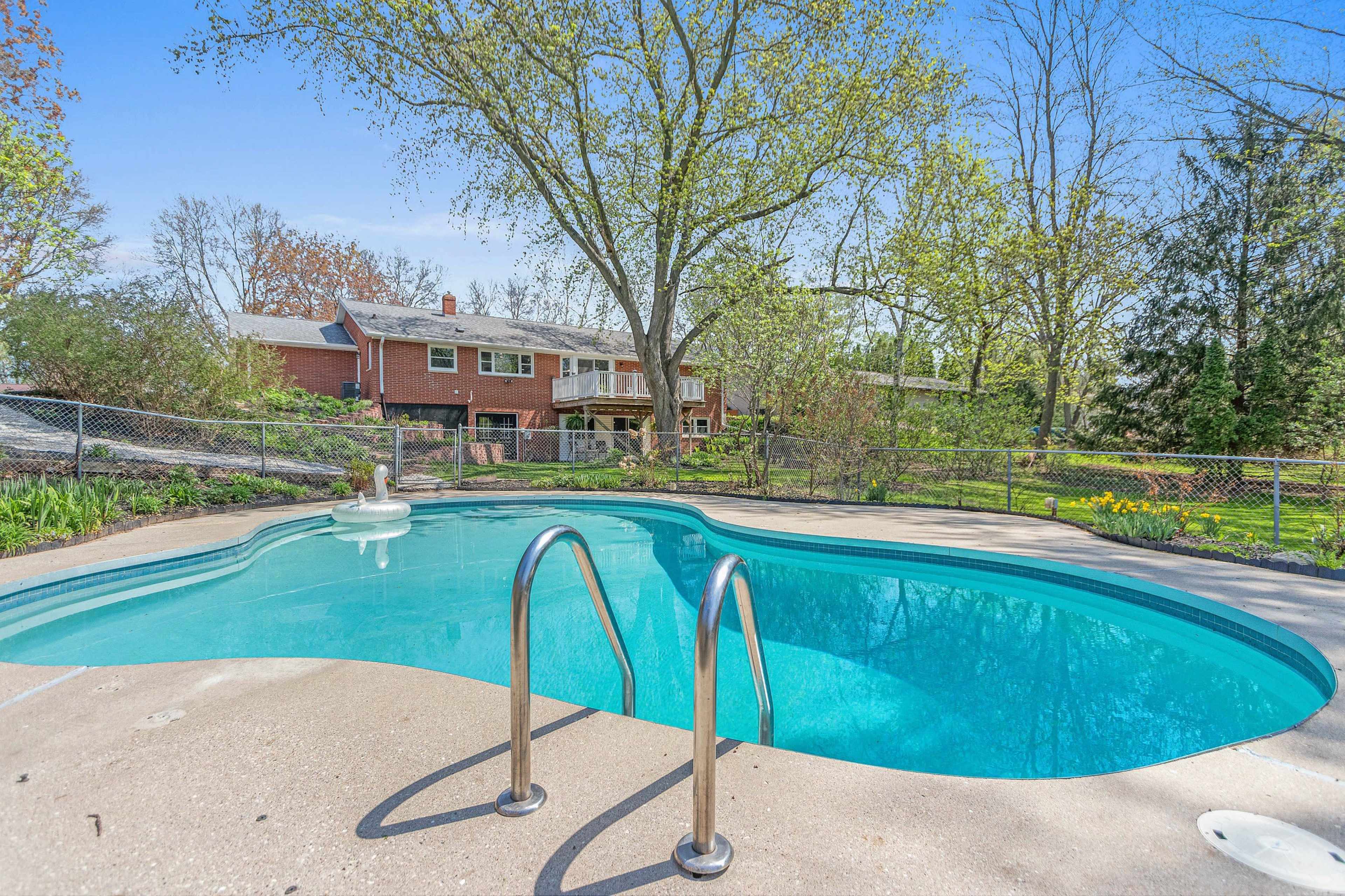 A swimming pool with a diving board is surrounded by a concrete deck and overlooks a large backyard with trees and a house in the background.