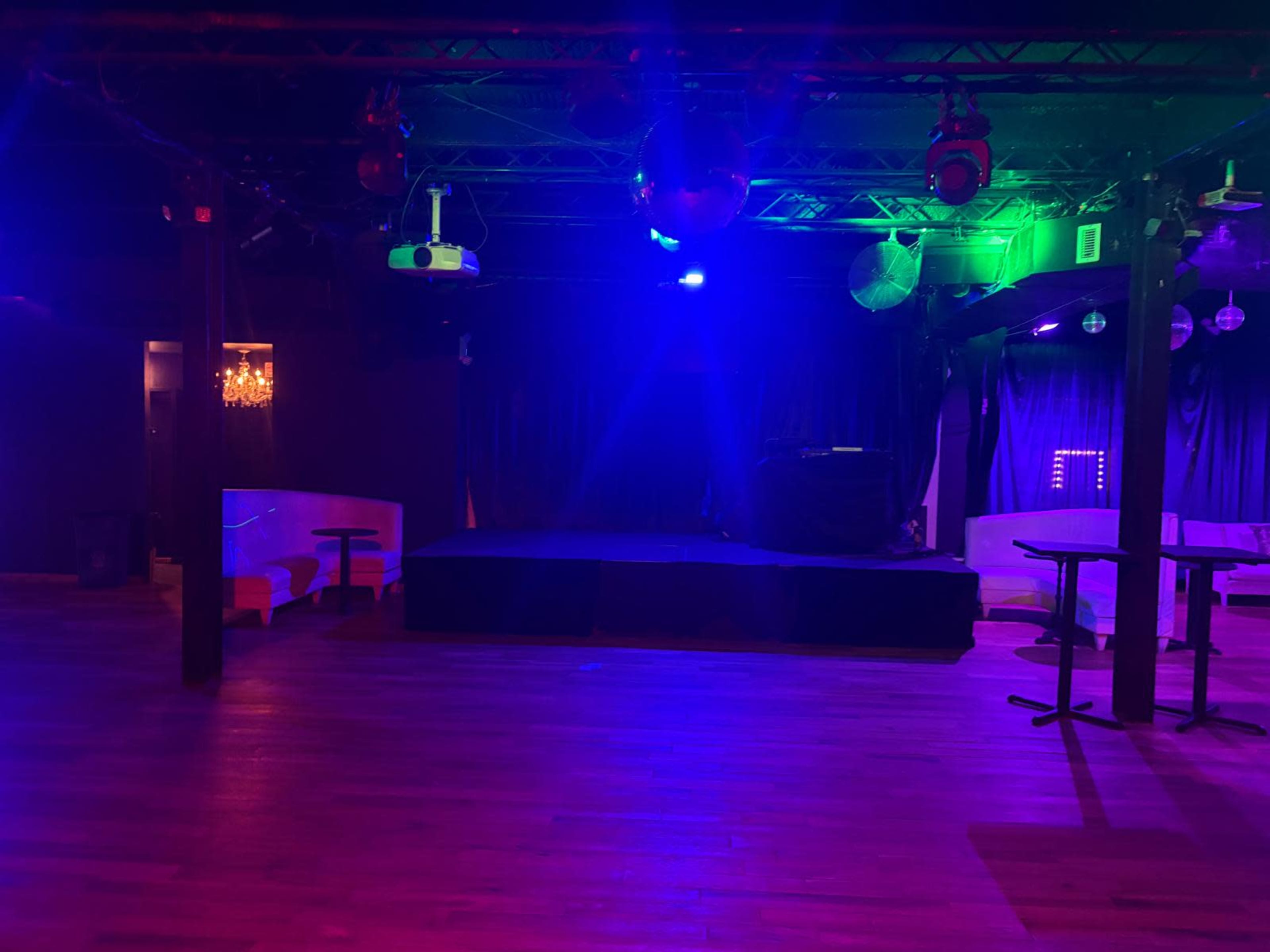 The image shows the interior of a dimly lit venue with a stage, seating areas, and colorful lighting effects.