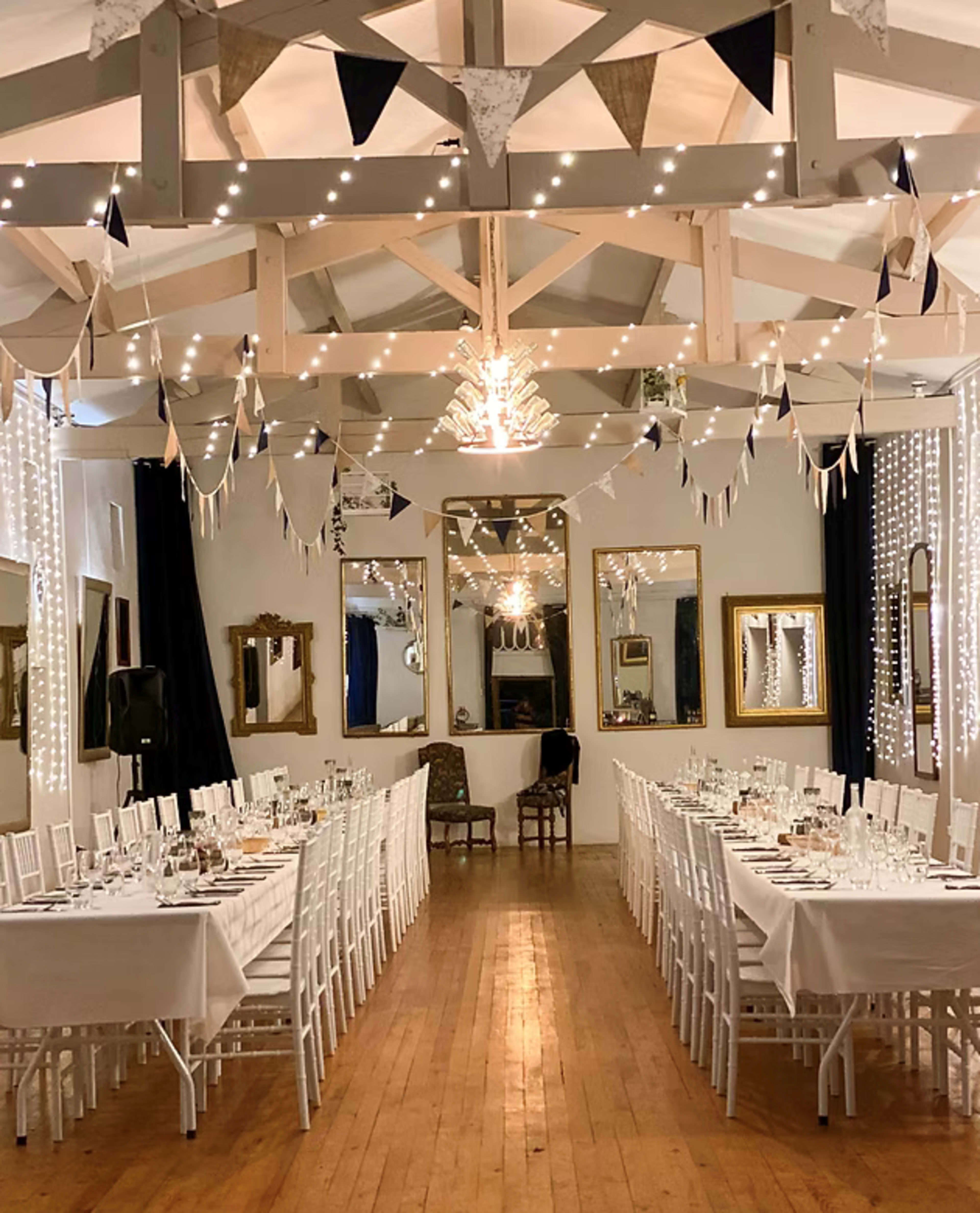 The image shows a decorated indoor venue with two long tables set for dining, arranged beneath twinkling lights and triangular bunting along the ceiling.