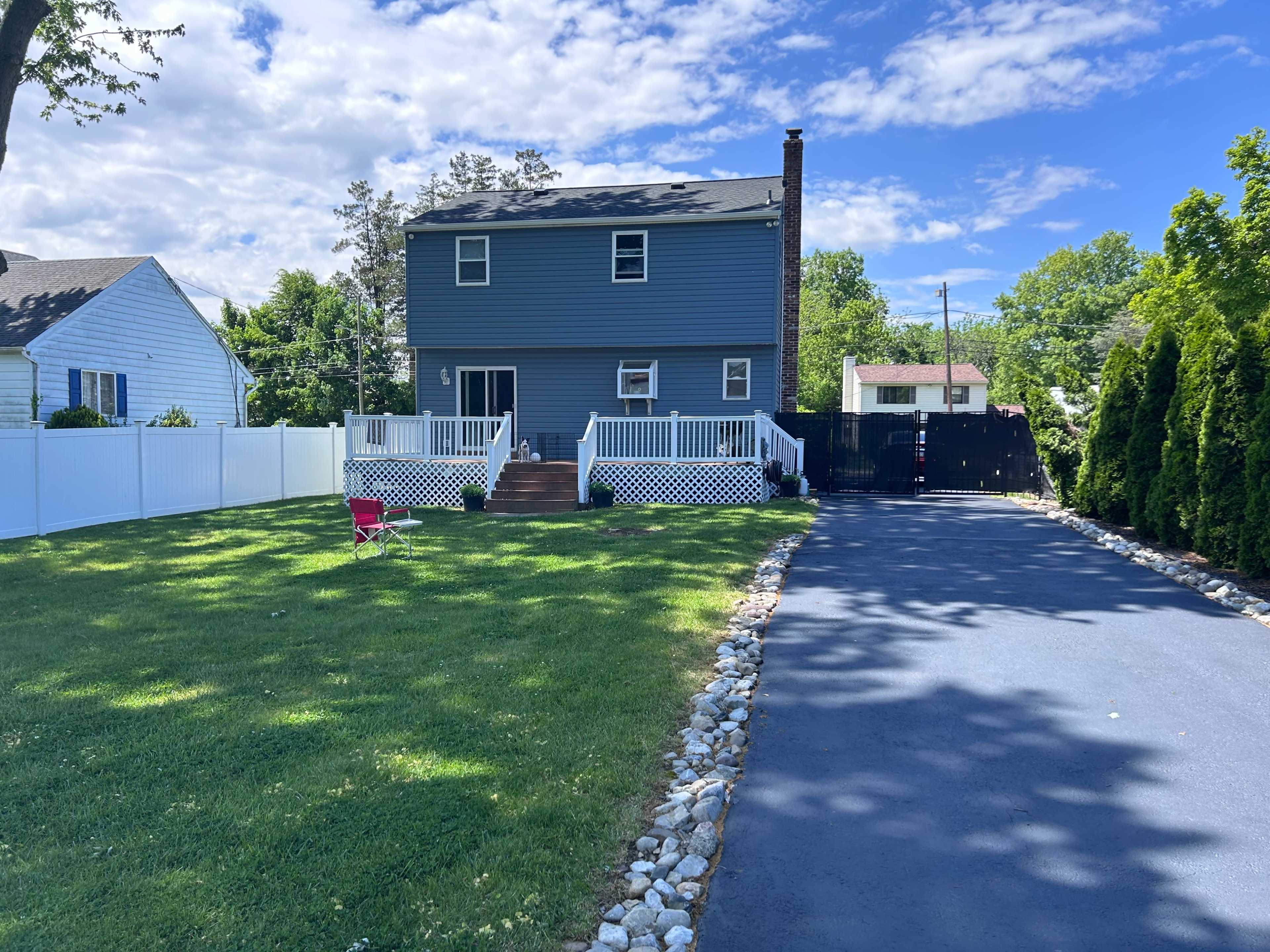 A blue two-story house with a white railing porch sits on a grassy lawn, bordered by a black fence and a paved driveway.