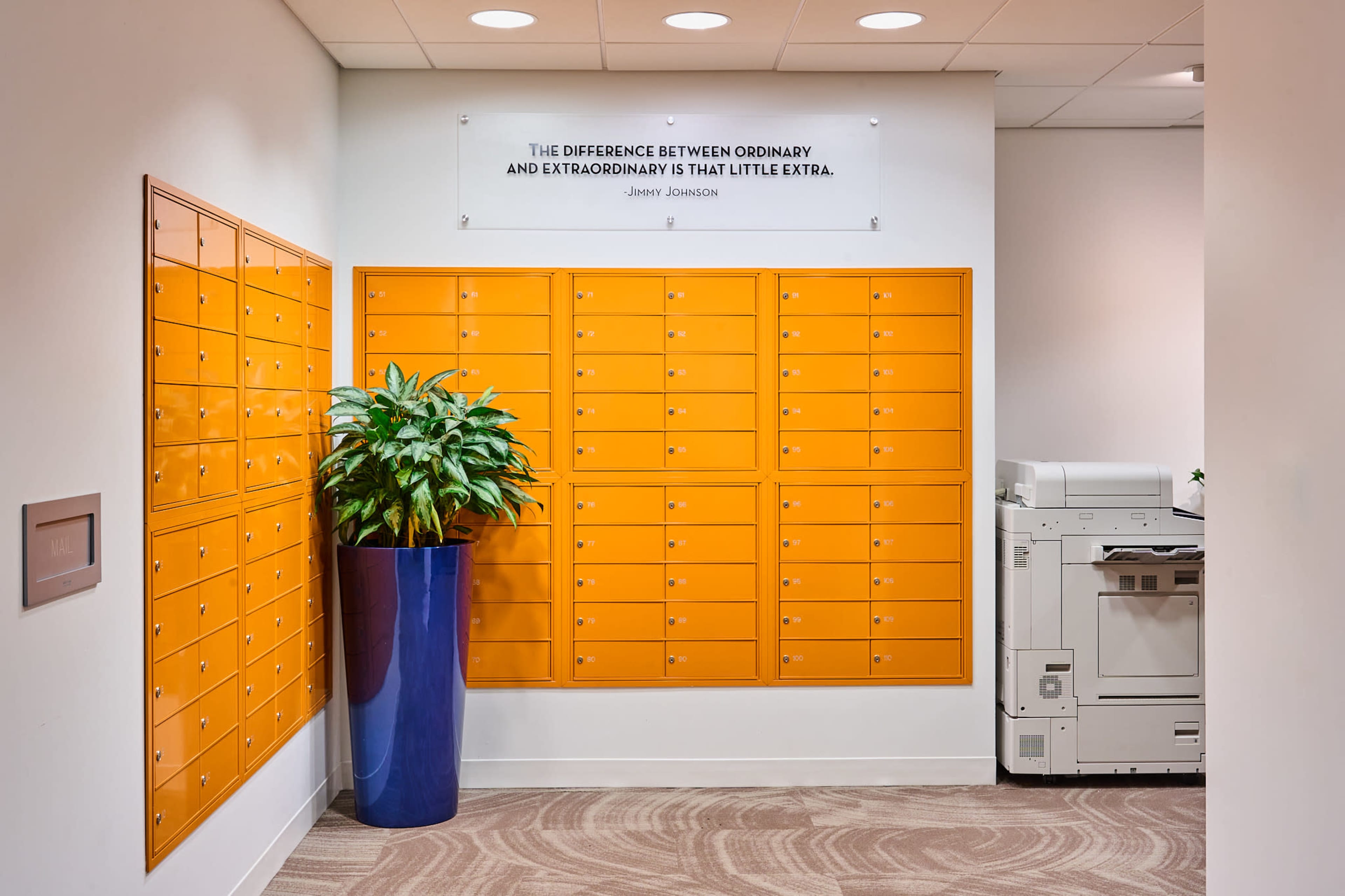The image shows a modern office space featuring a wall of orange mailboxes, a tall potted plant, and a printer near the entrance.