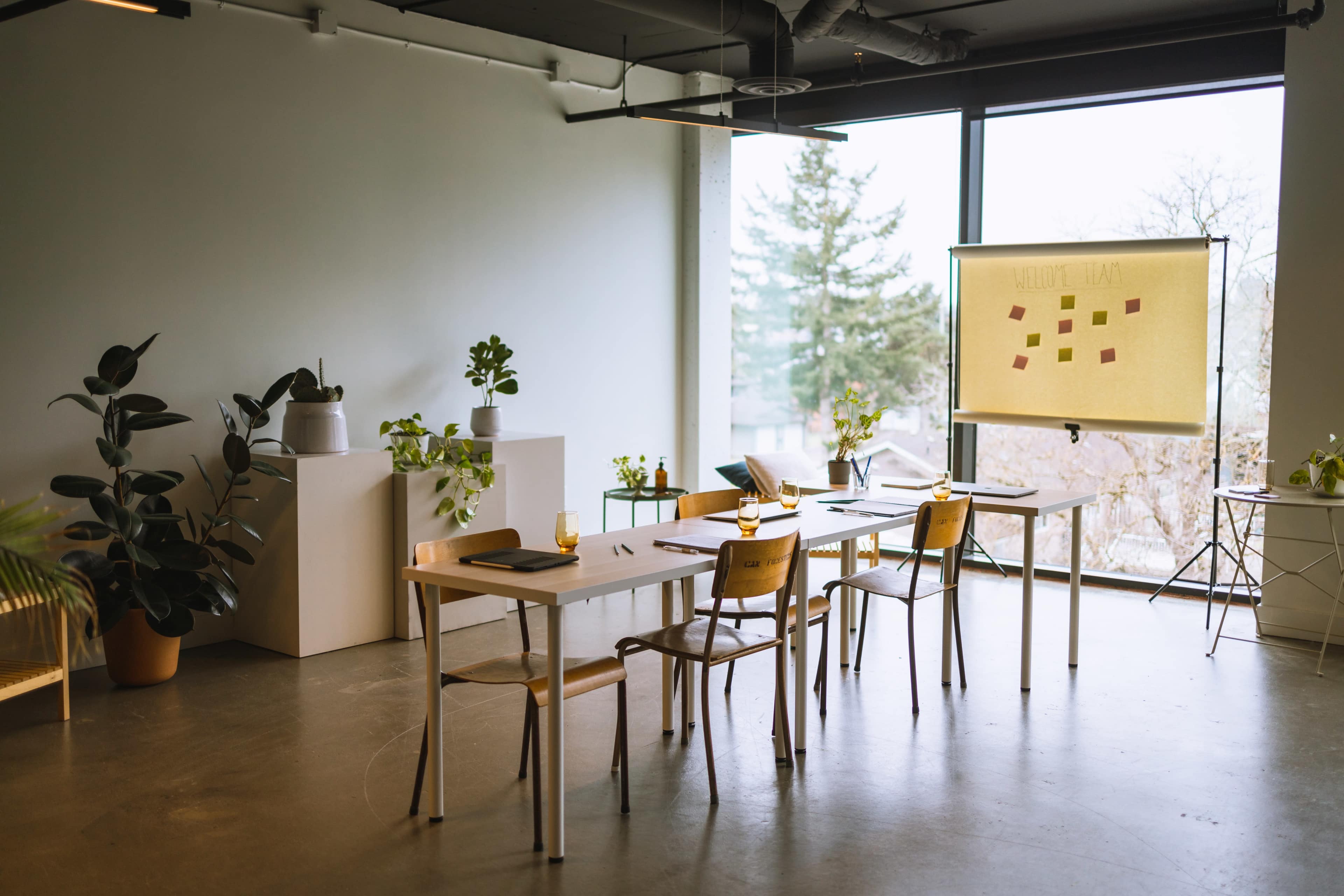 A bright meeting room features a long table with chairs, a projector screen displaying notes, and several potted plants near large windows.