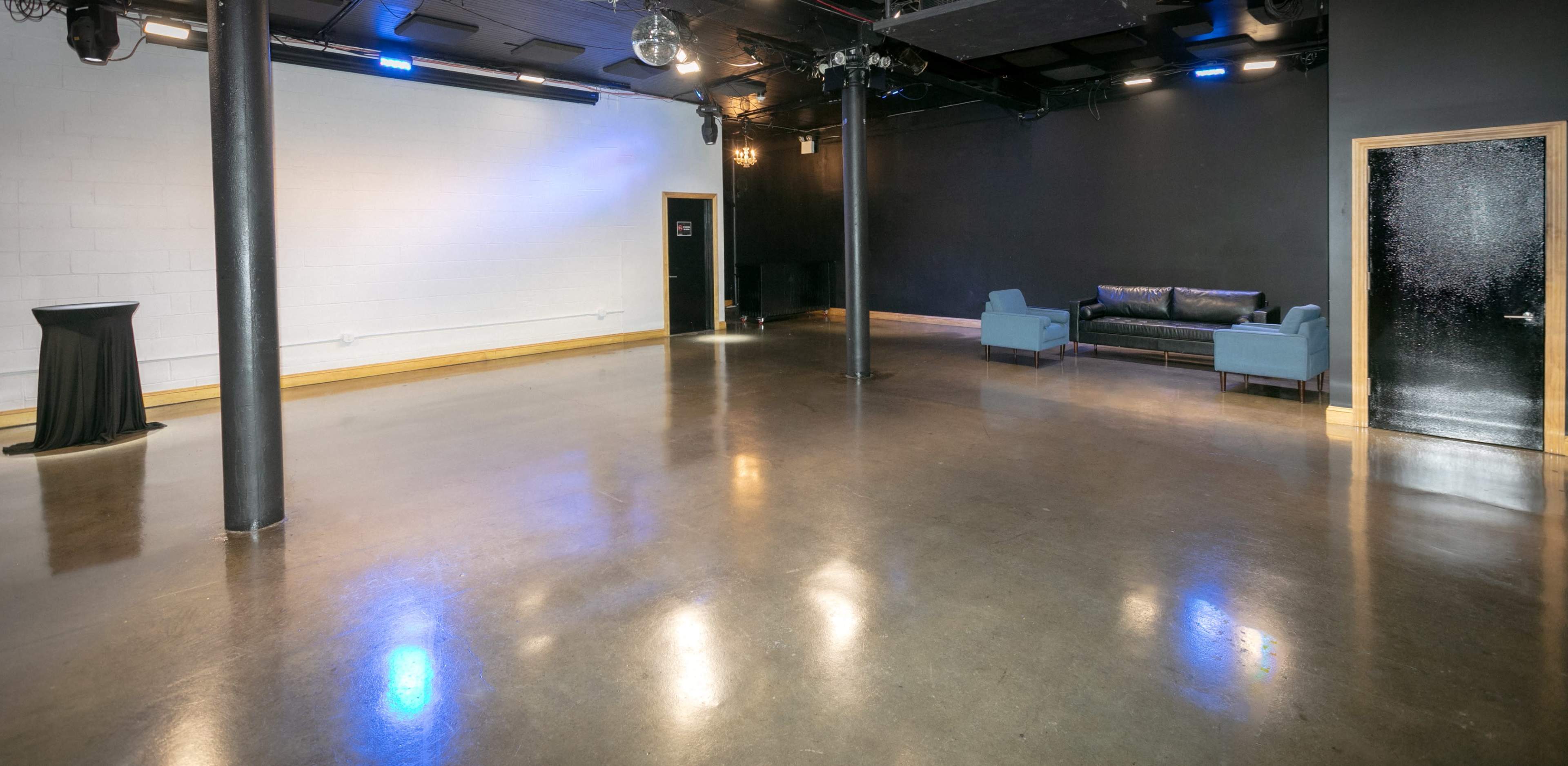 The image shows a spacious, mostly empty room with polished concrete floors, a black wall, a blue couch, and a tall black table in one corner.