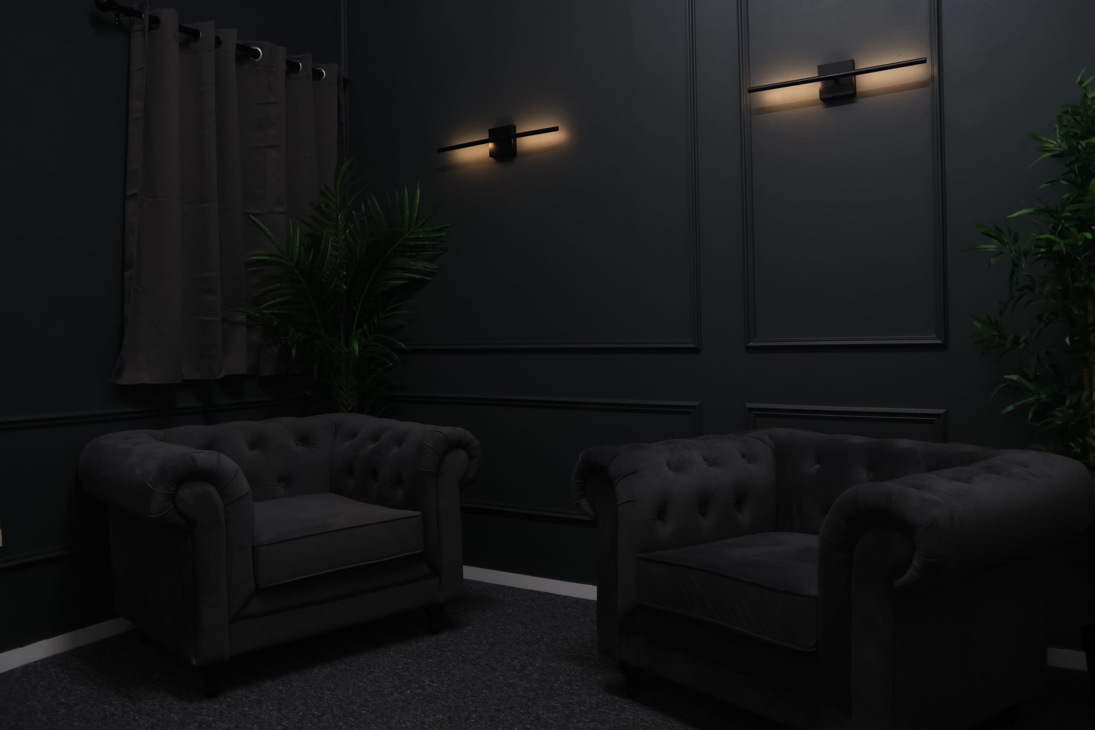 The image shows two dark upholstered armchairs positioned in a dimly lit room with black walls and modern wall sconces, complemented by a potted plant.