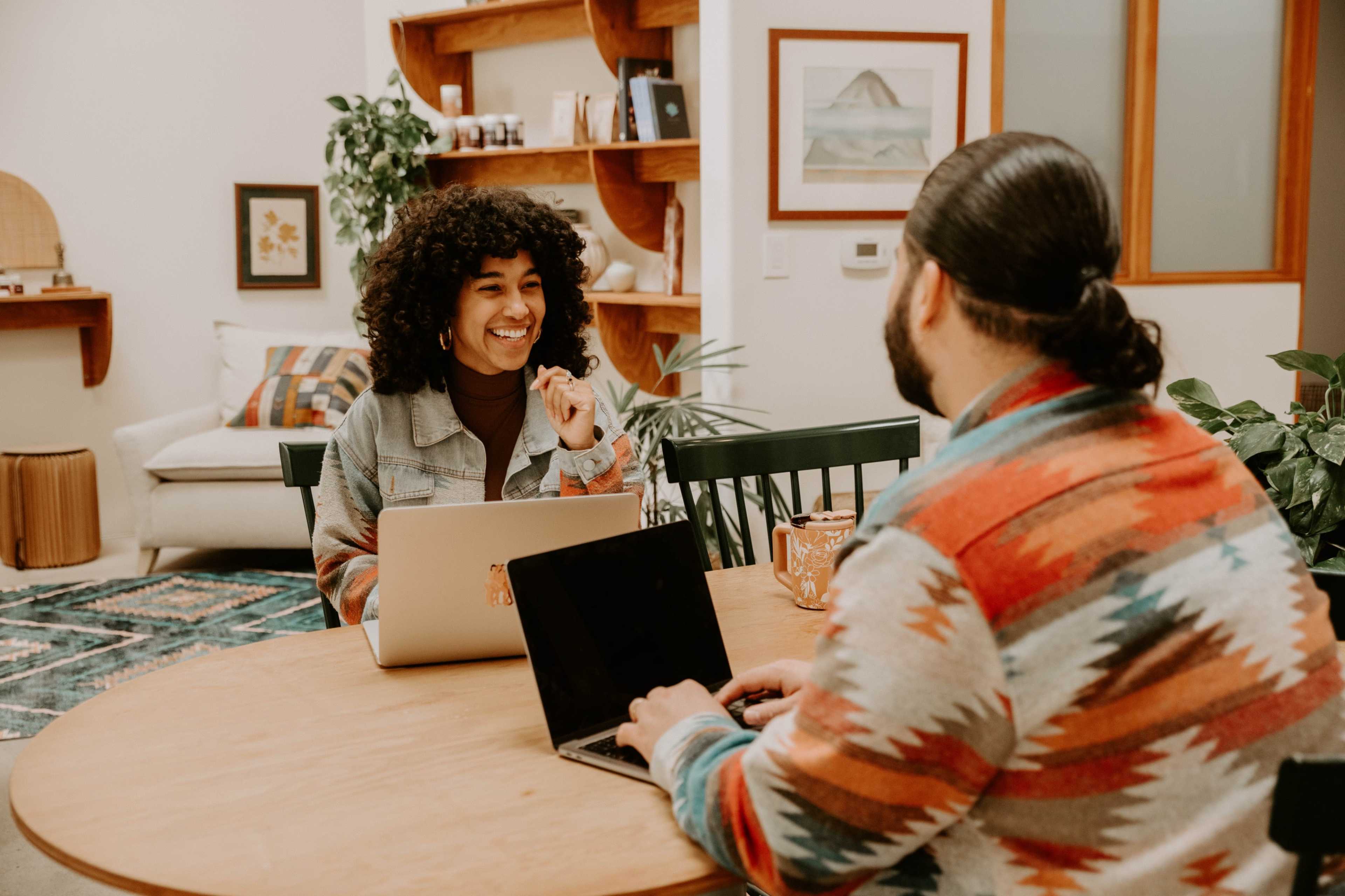 A woman with curly hair and a man with long hair are seated at a wooden table, each working on their laptops in a well-lit, cozy room decorated with plants and wall art.