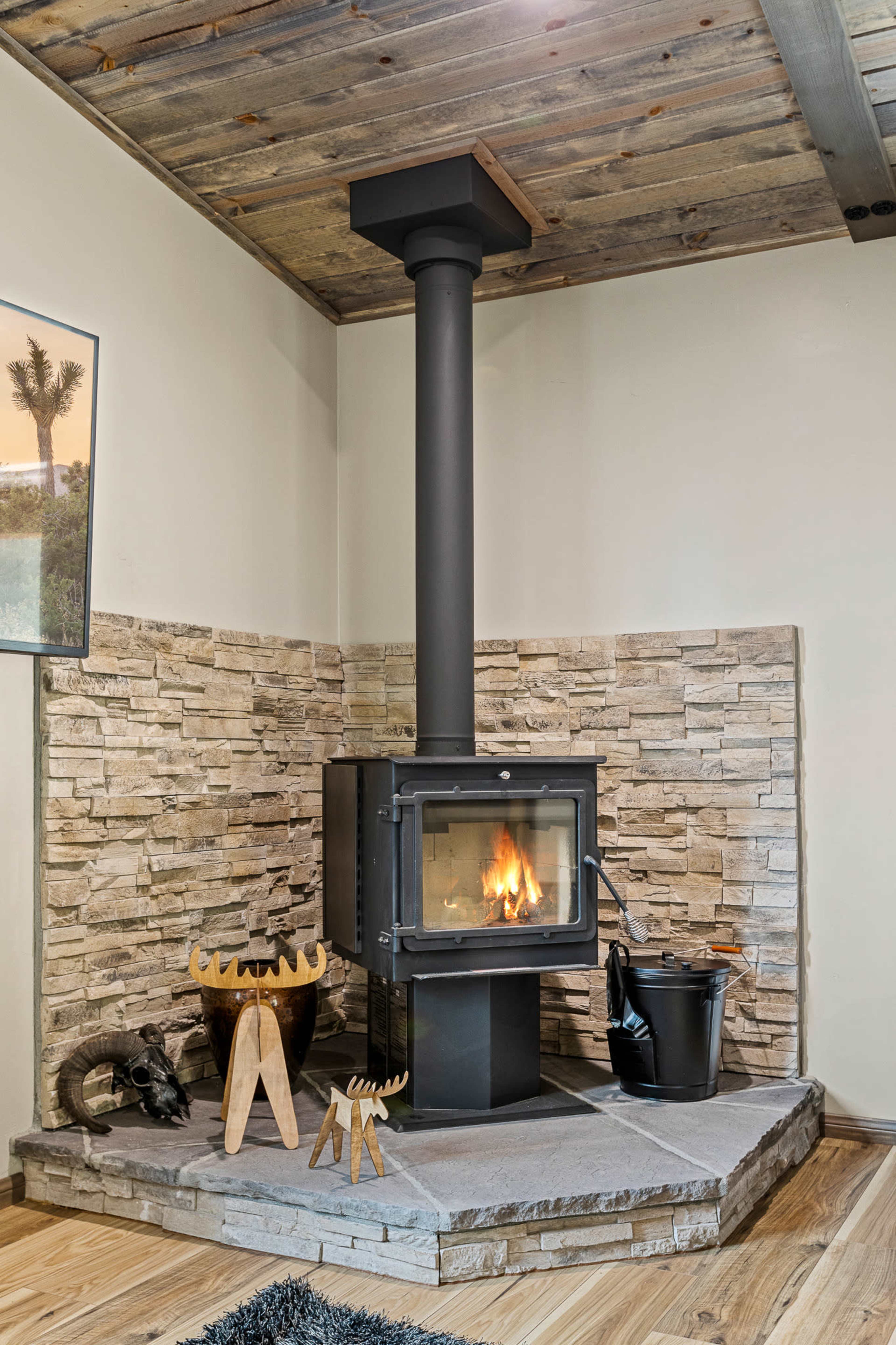 A modern wood stove with a flue pipe is positioned in the corner of a room, surrounded by a stone wall and a slate base, with a few decorative wooden and metal accents nearby.