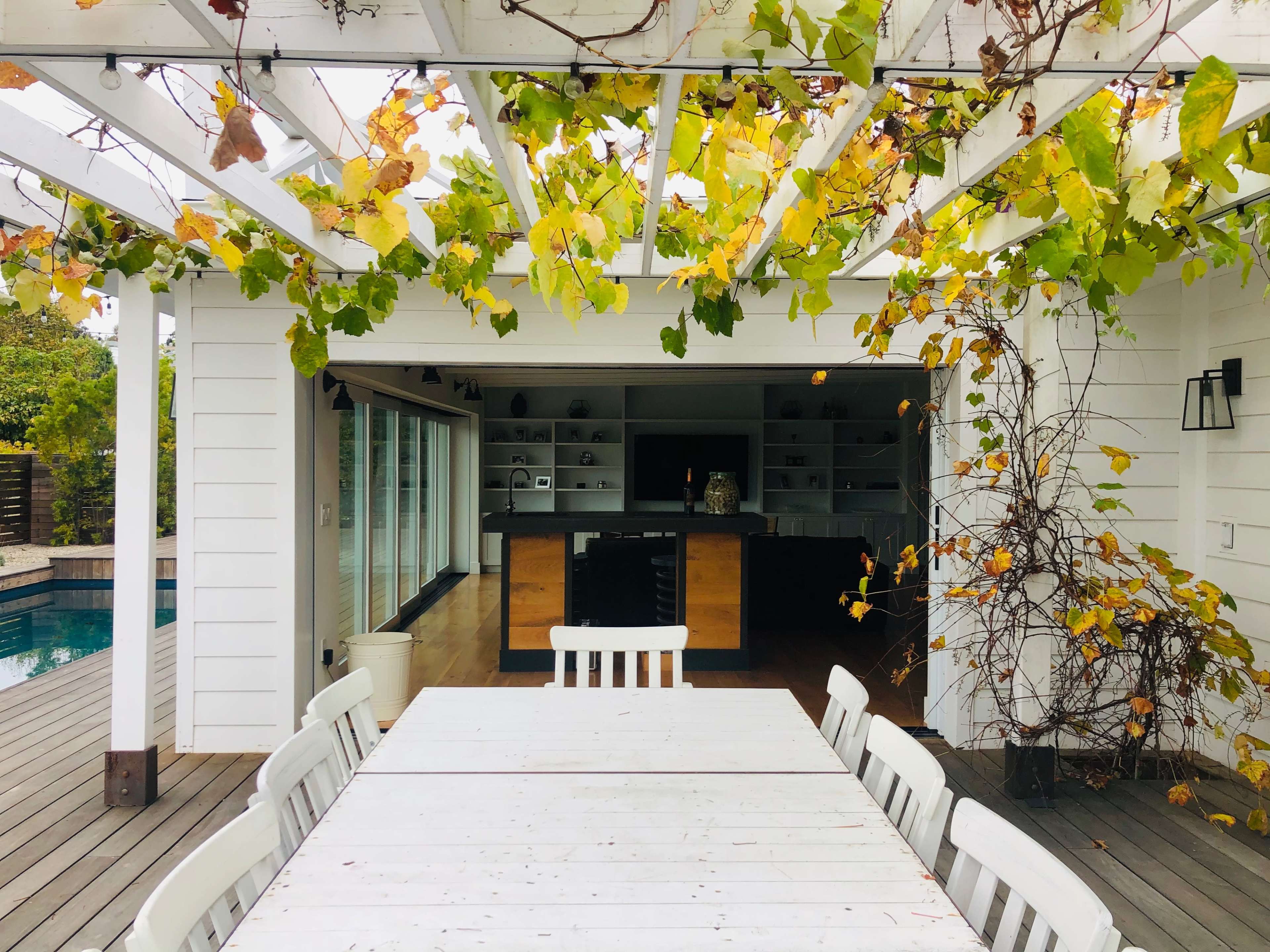 A white outdoor table is set under a pergola adorned with grapevines, overlooking a modern kitchen and a swimming pool.