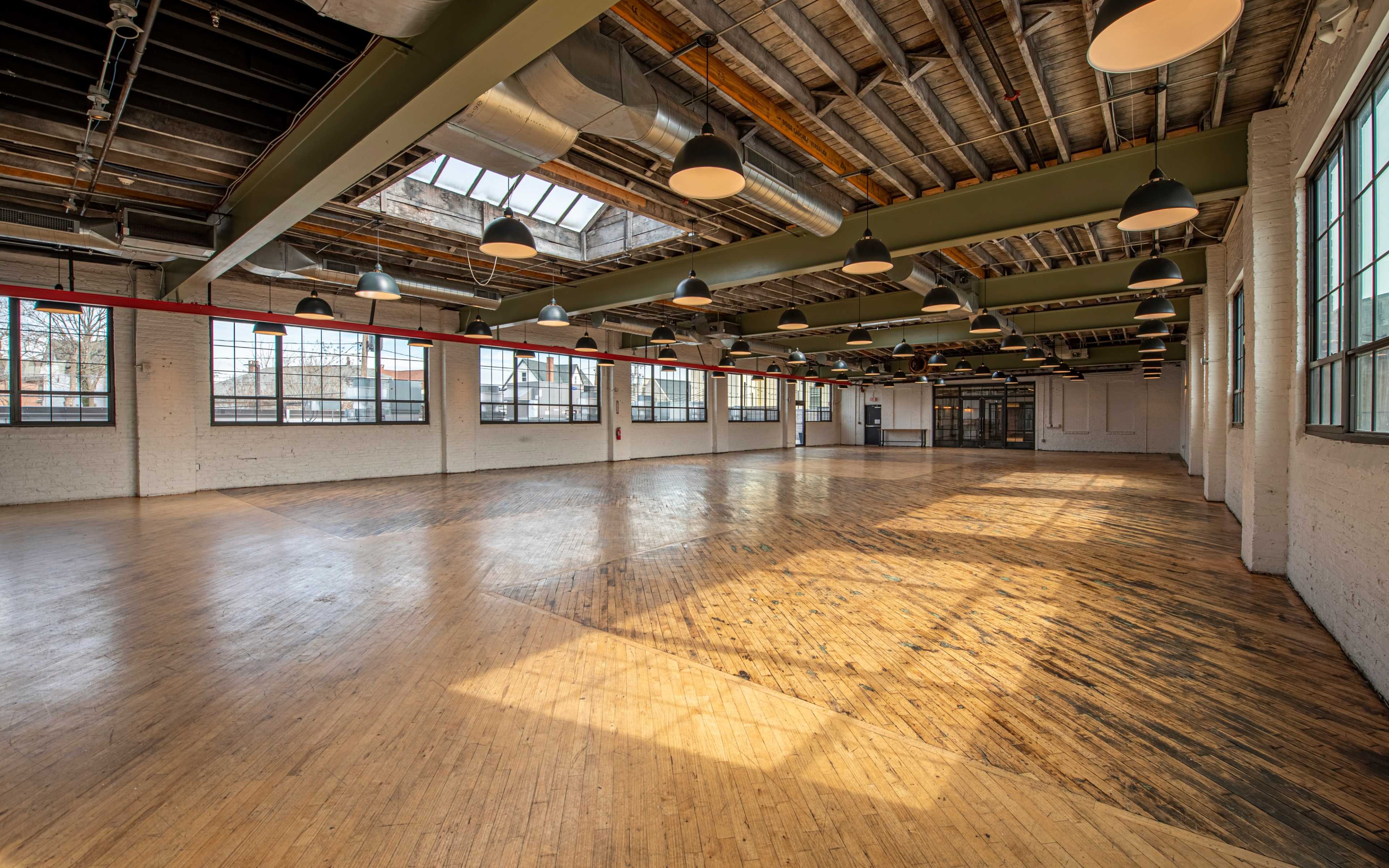 A spacious, empty indoor hall with wooden floors, large windows, and overhead lighting.