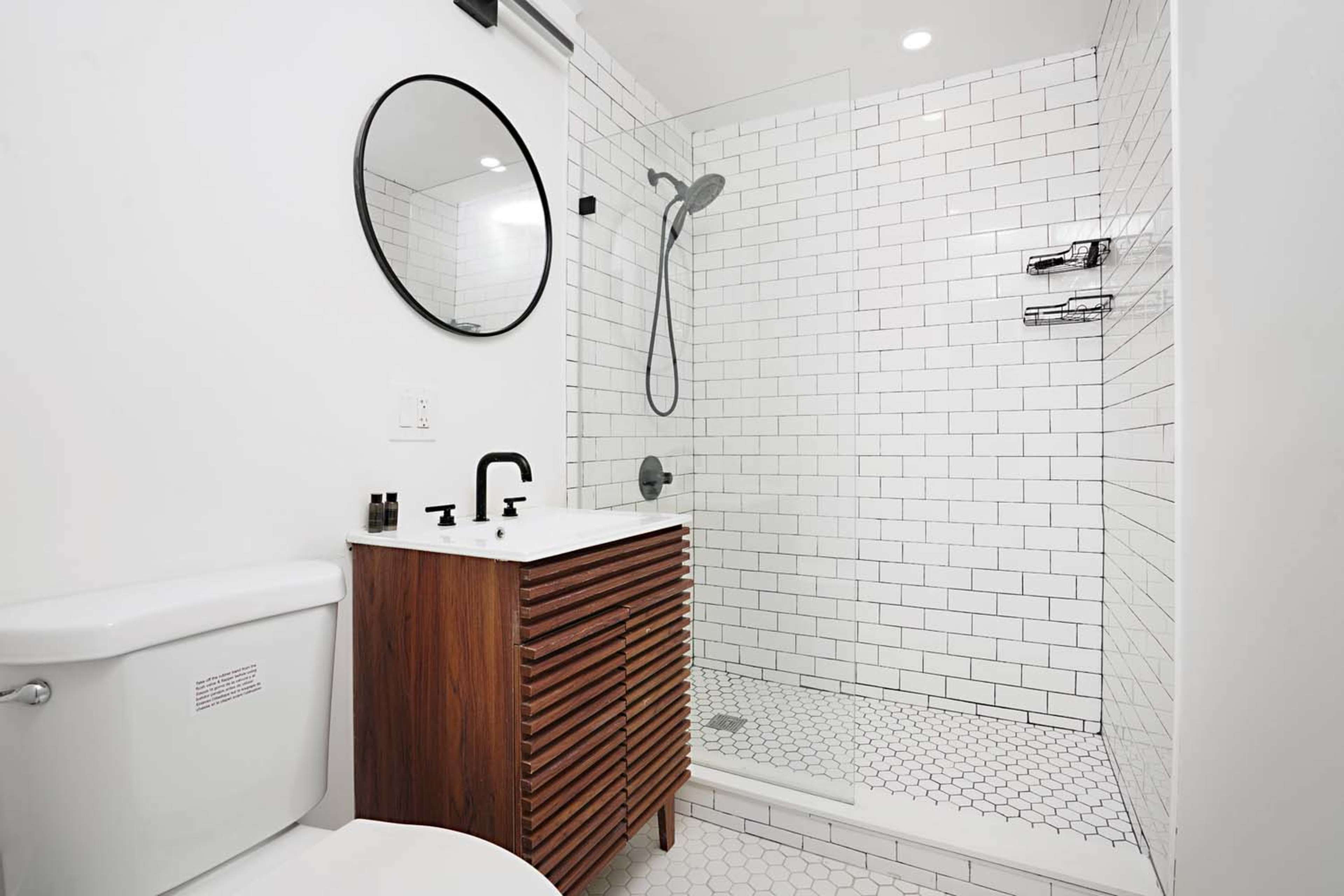 The image features a modern bathroom with a glass shower enclosure, a round mirror, a wooden vanity, and white tiled walls and floor.