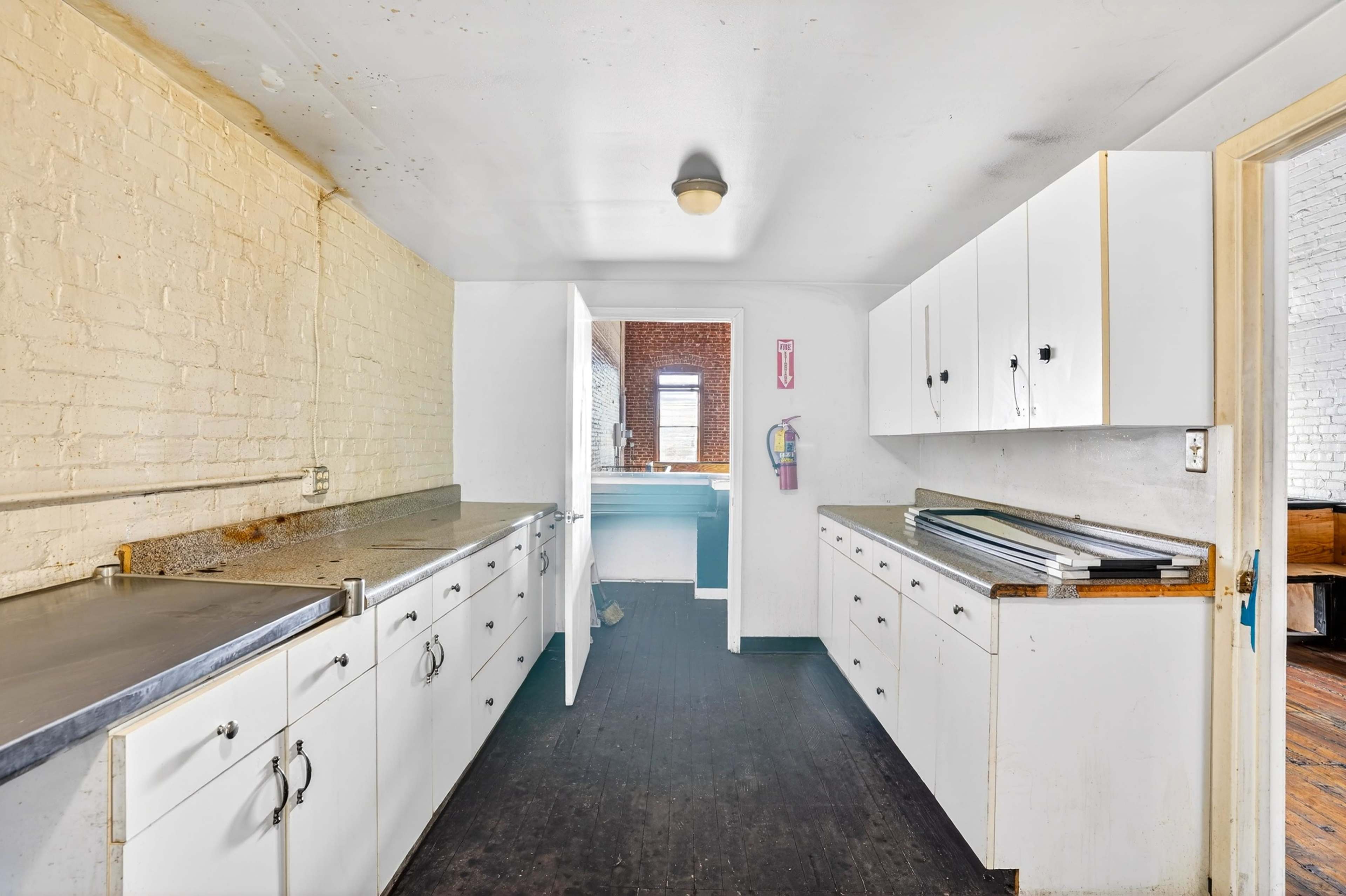 The image shows a narrow kitchen with white cabinets and stainless steel countertops, featuring a door leading to another room at the far end.