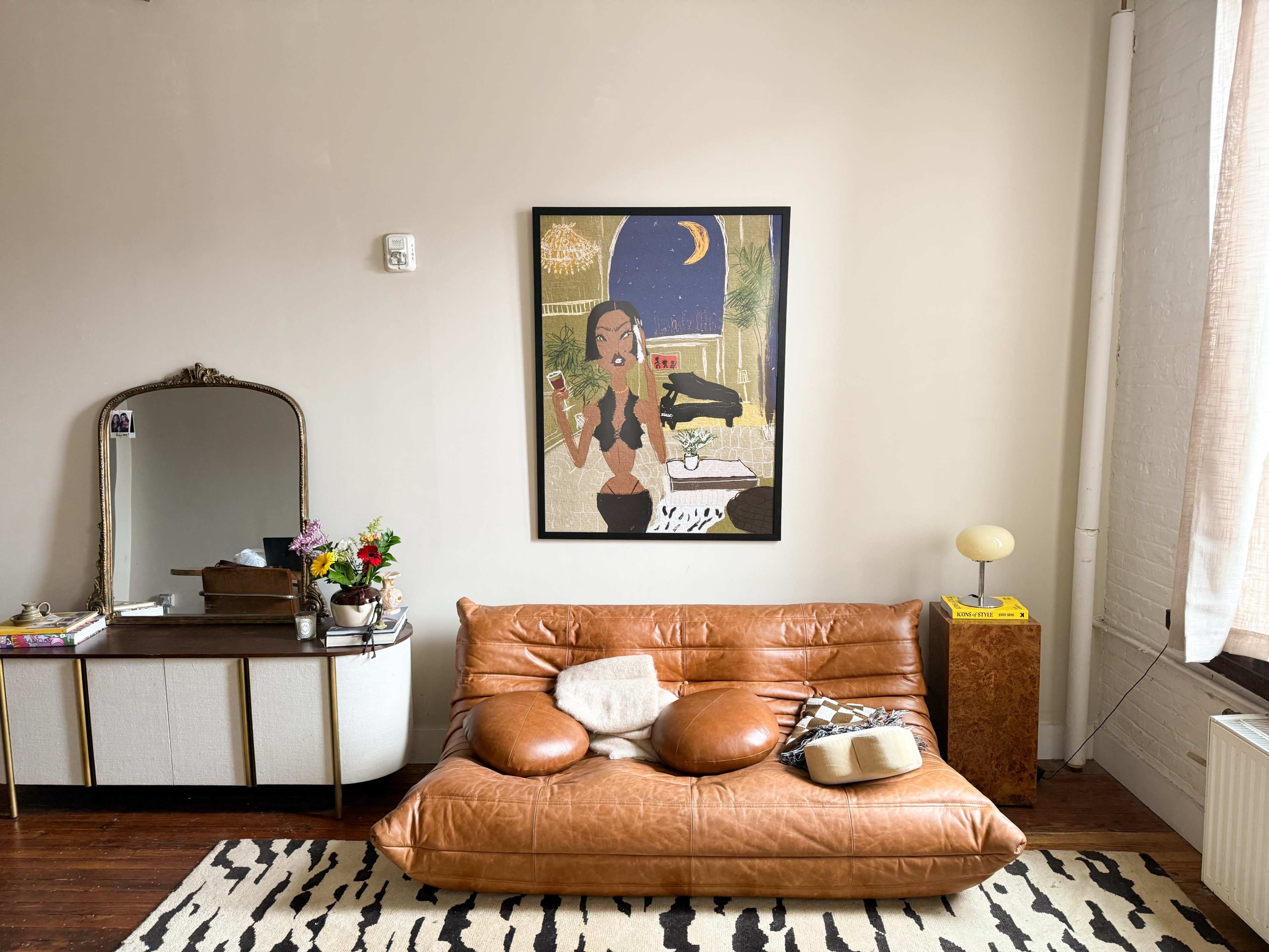 The image shows a contemporary interior with a brown leather sofa, an art piece featuring a woman, and a decorative mirror on a side table.