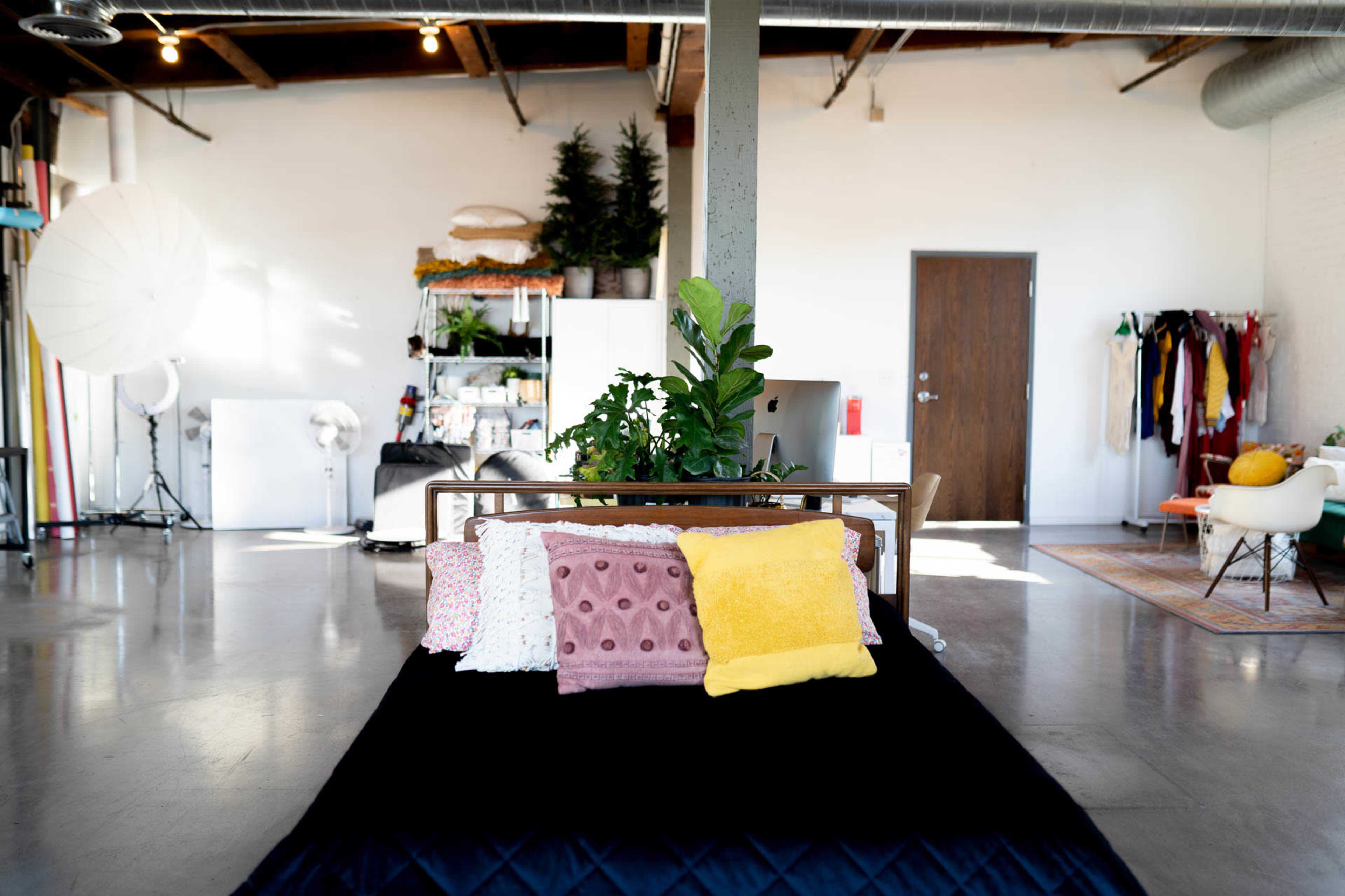 A minimalist bedroom features a bed with colorful pillows, a potted plant, and an open space that includes a clothing rack and various furnishings.