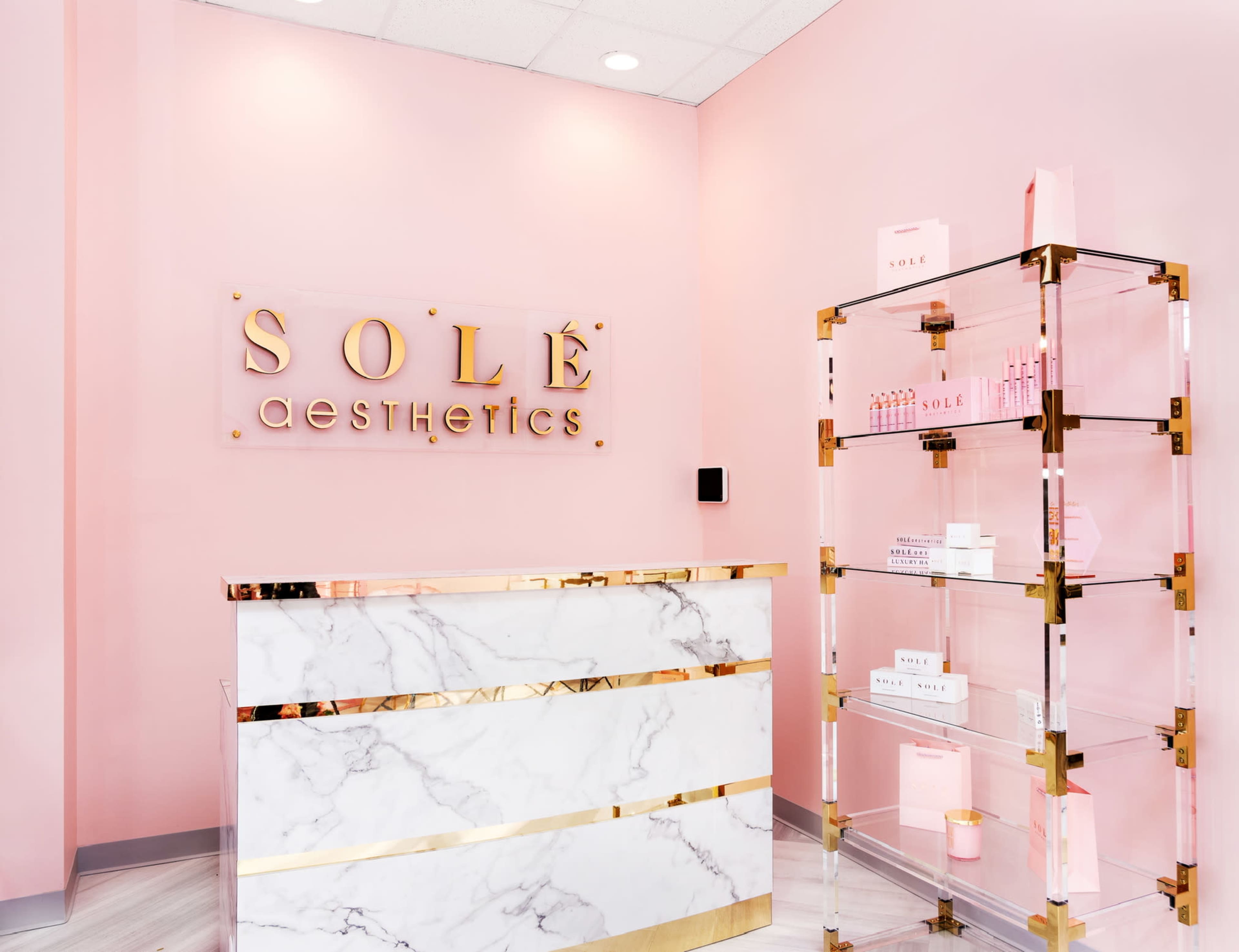 The image shows a modern aesthetics clinic reception area with a pink wall, a marble front desk, and a transparent display shelf holding skincare products.