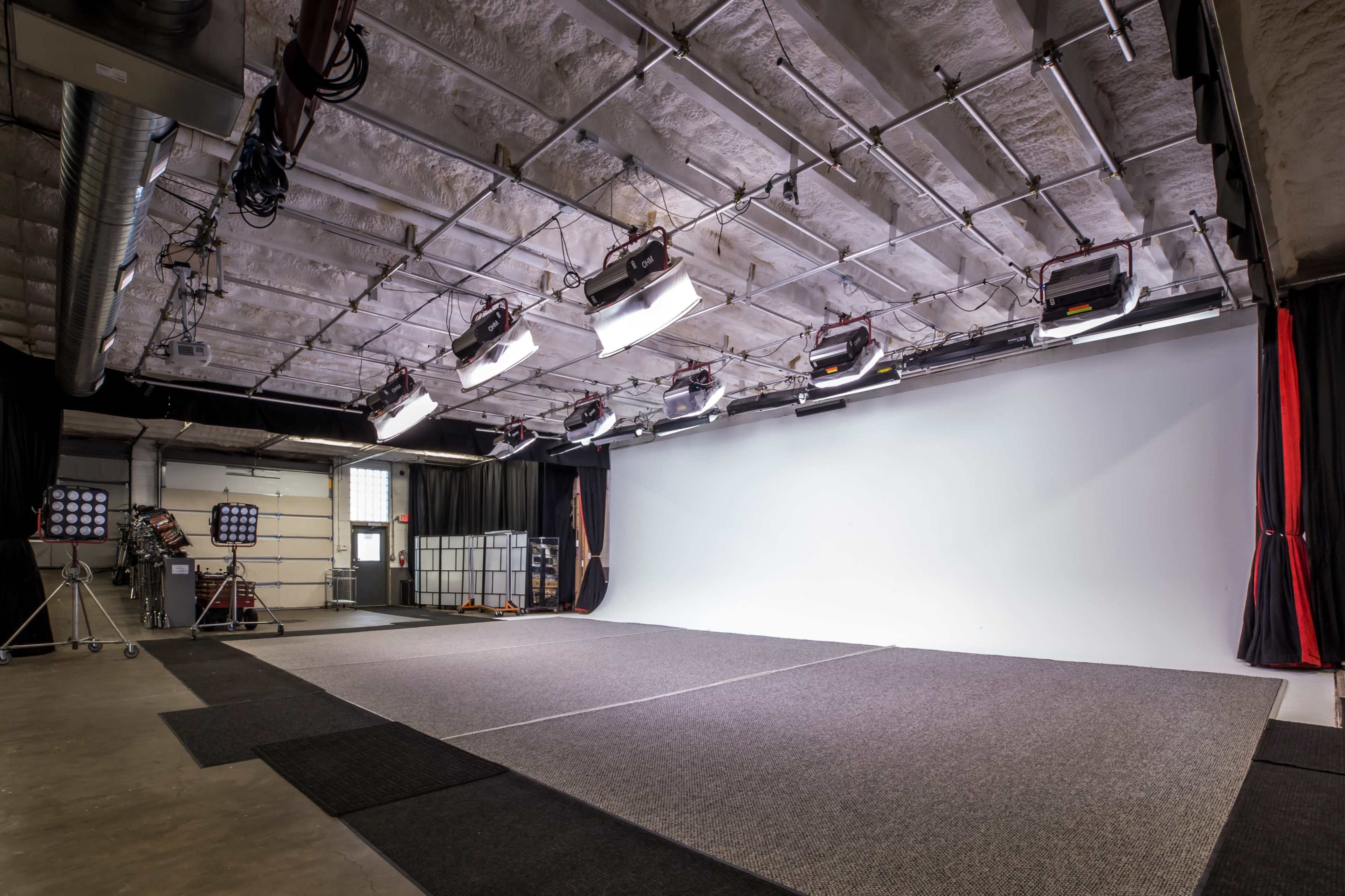A photography studio with a large white backdrop, multiple lights mounted on the ceiling, and dark curtains along the sides.