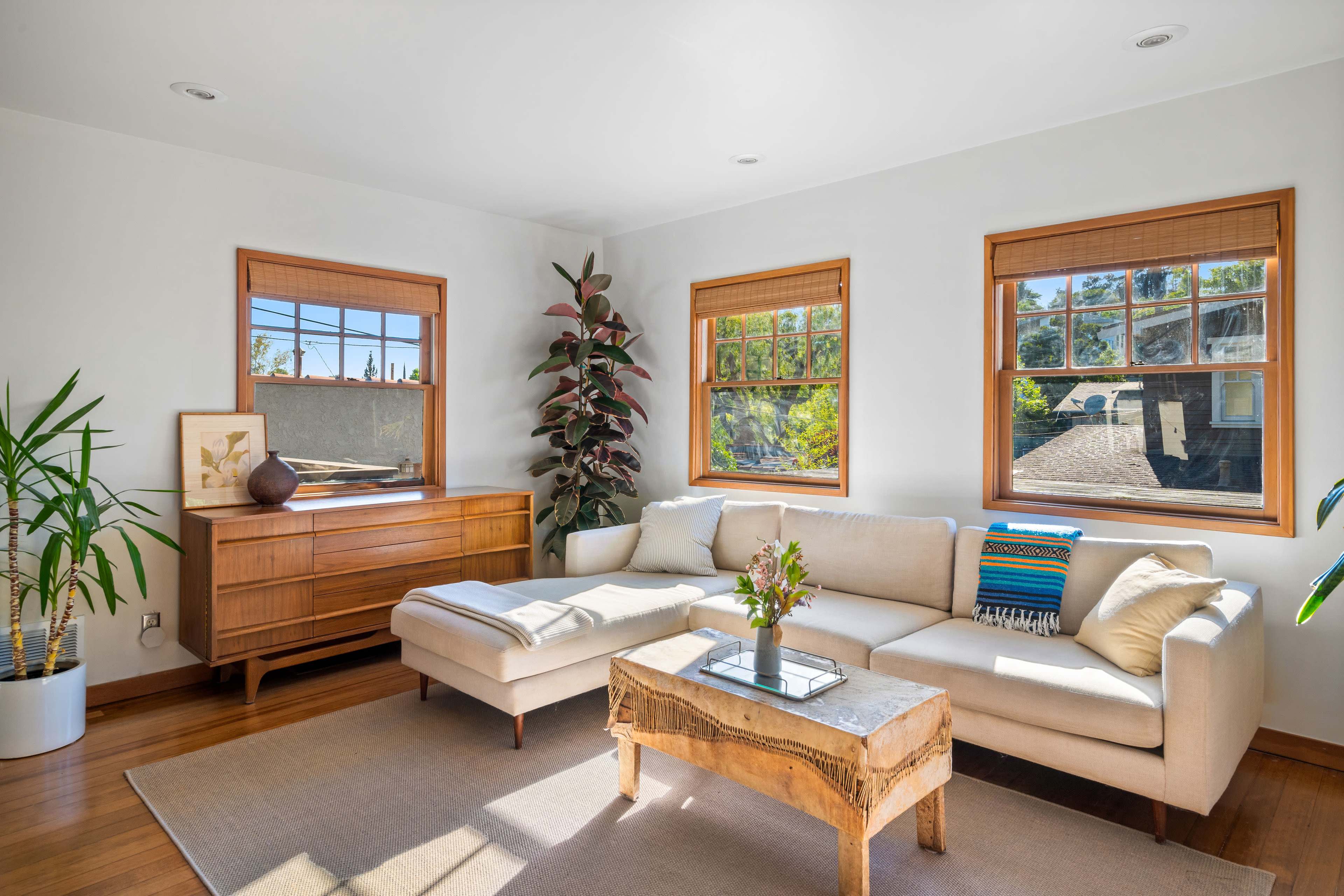 A light-filled living room features a beige sectional sofa, a wooden coffee table, and large windows decorated with bamboo shades.