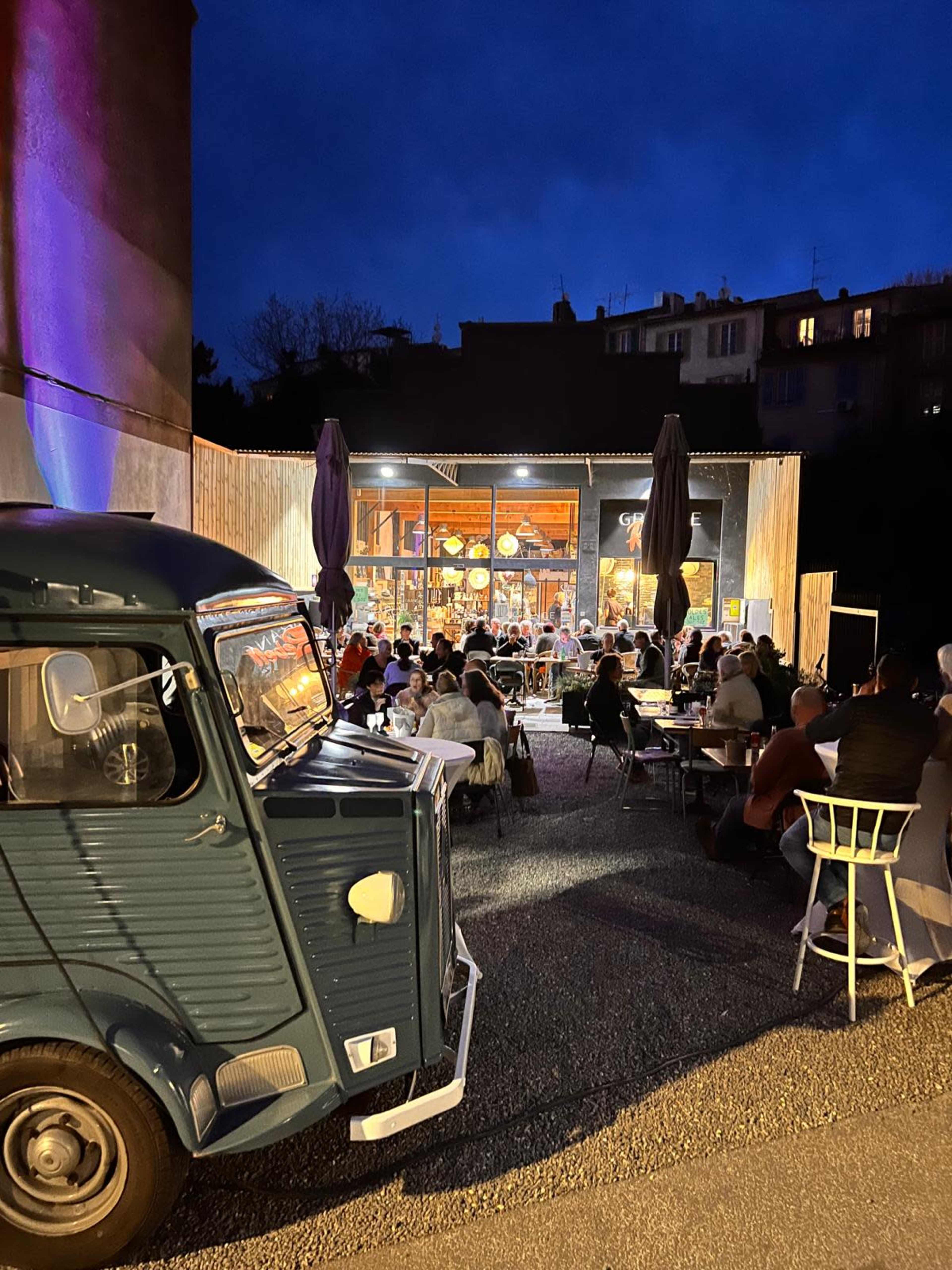 A vintage blue truck is parked in front of a restaurant with outdoor seating, where patrons are dining under evening lights.