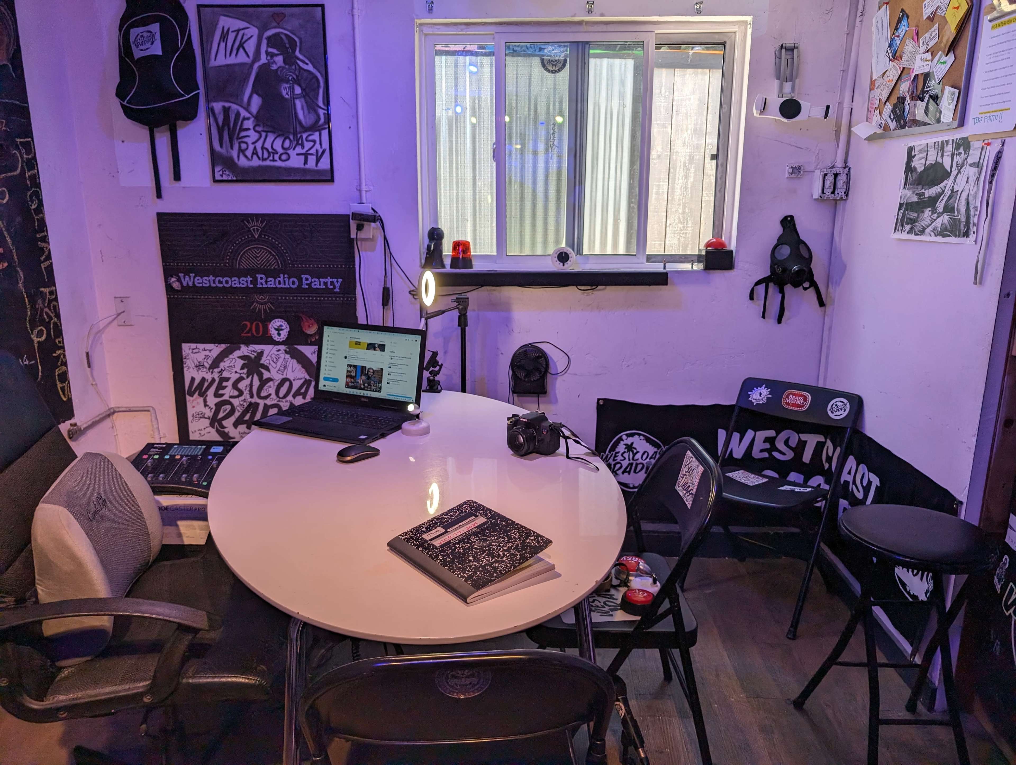 A small room features a round table with a laptop, a gaming console, and a book, surrounded by black folding chairs and decorated with music-themed posters.