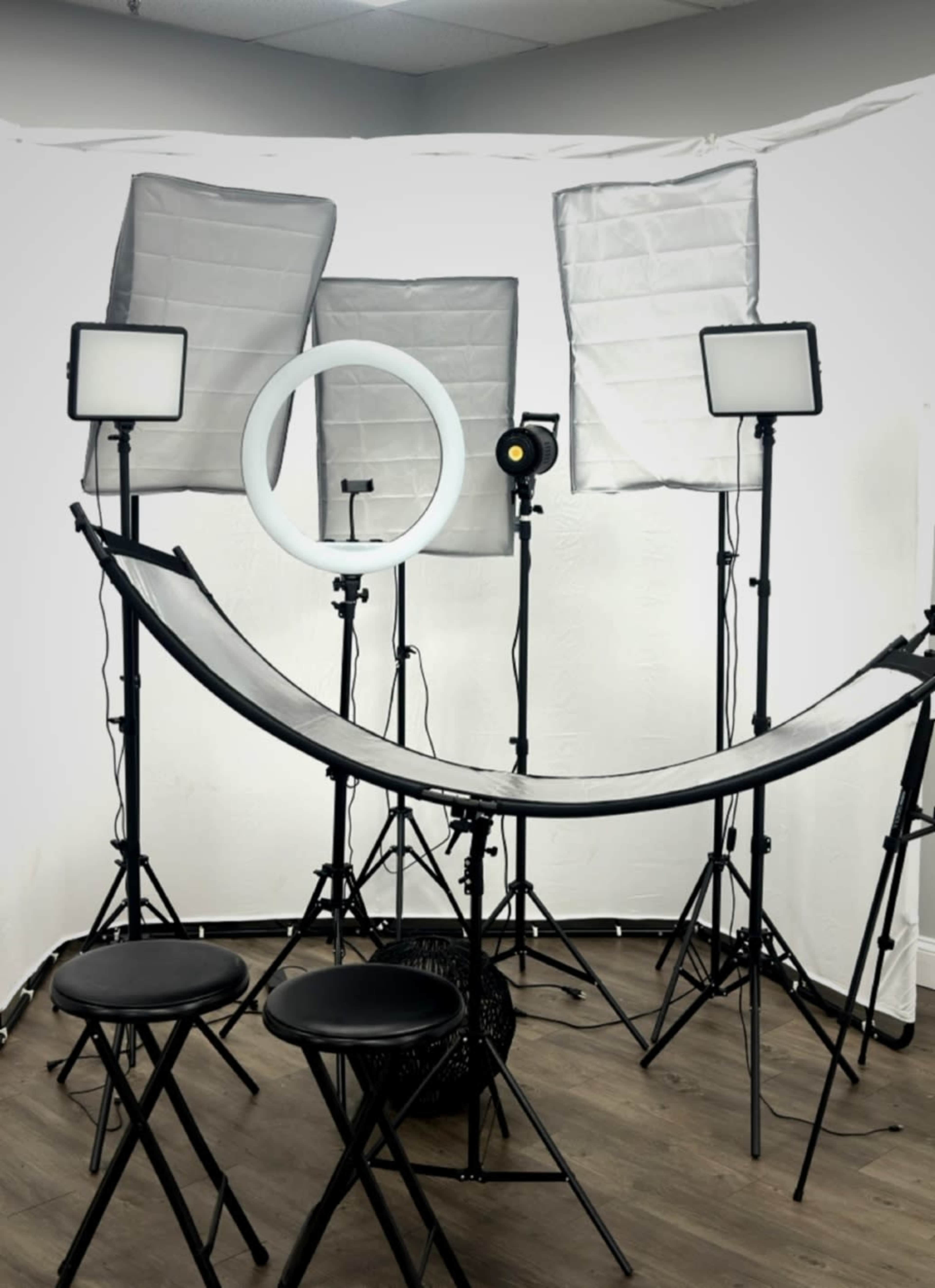 The image shows a photography setup with multiple softbox lights, a ring light, and two black stools arranged in a studio space.