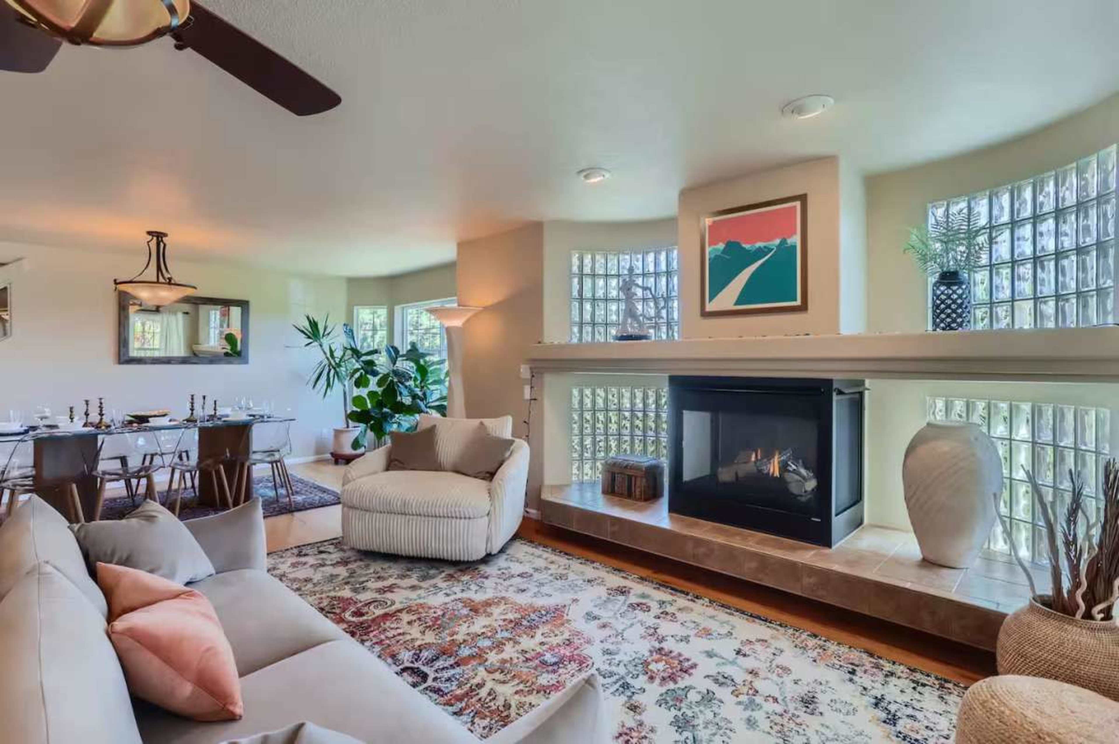 The image shows a modern living room featuring a cozy seating area, a fireplace, a large rug, and decorative plants near a wall with glass block windows.
